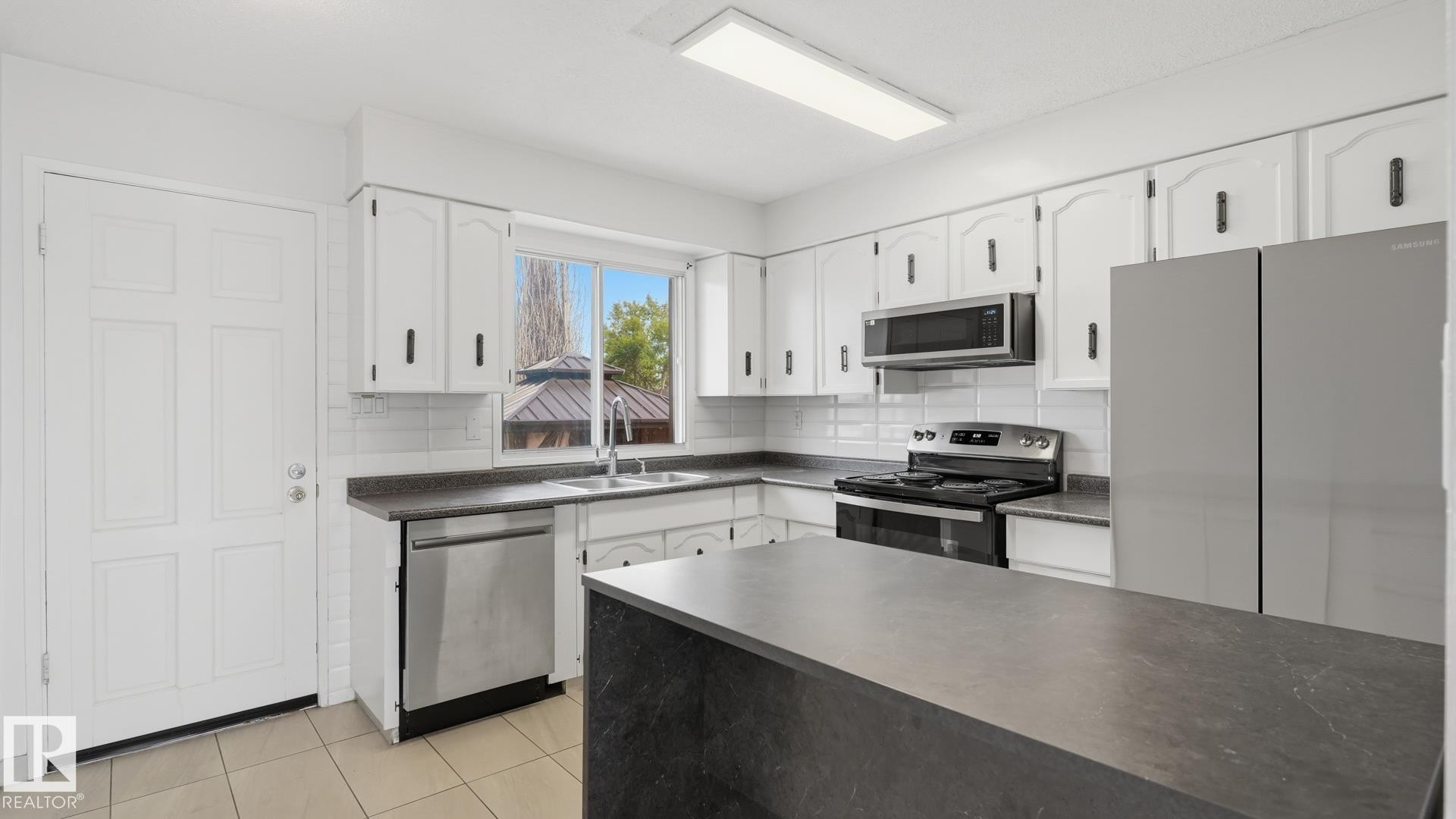 White cabinetry with black hardware, stainless steel appliances, dark countertops, a kitchen island, and tiled flooring - 12208 146 Avenue Nw, Edmonton, AB