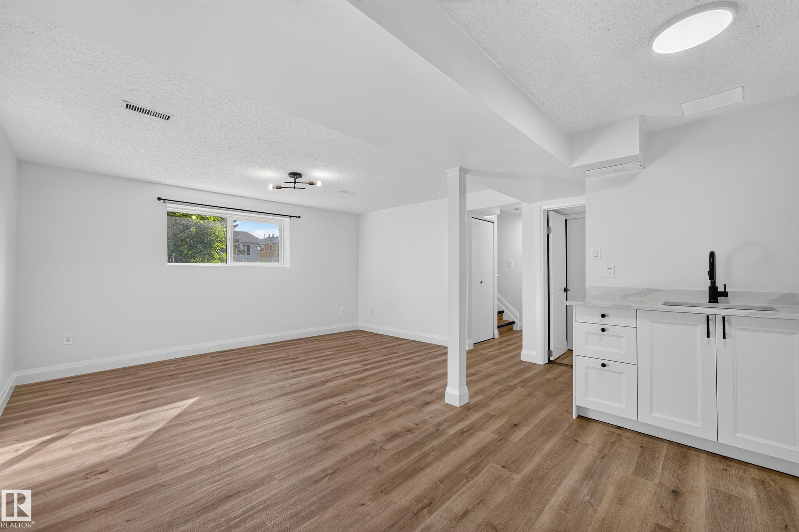 Spacious lower-level room featuring wood-finish flooring, a window, and a wet bar with white cabinetry, a countertop, and a black faucet - 2412 80 Street Nw, Edmonton, AB