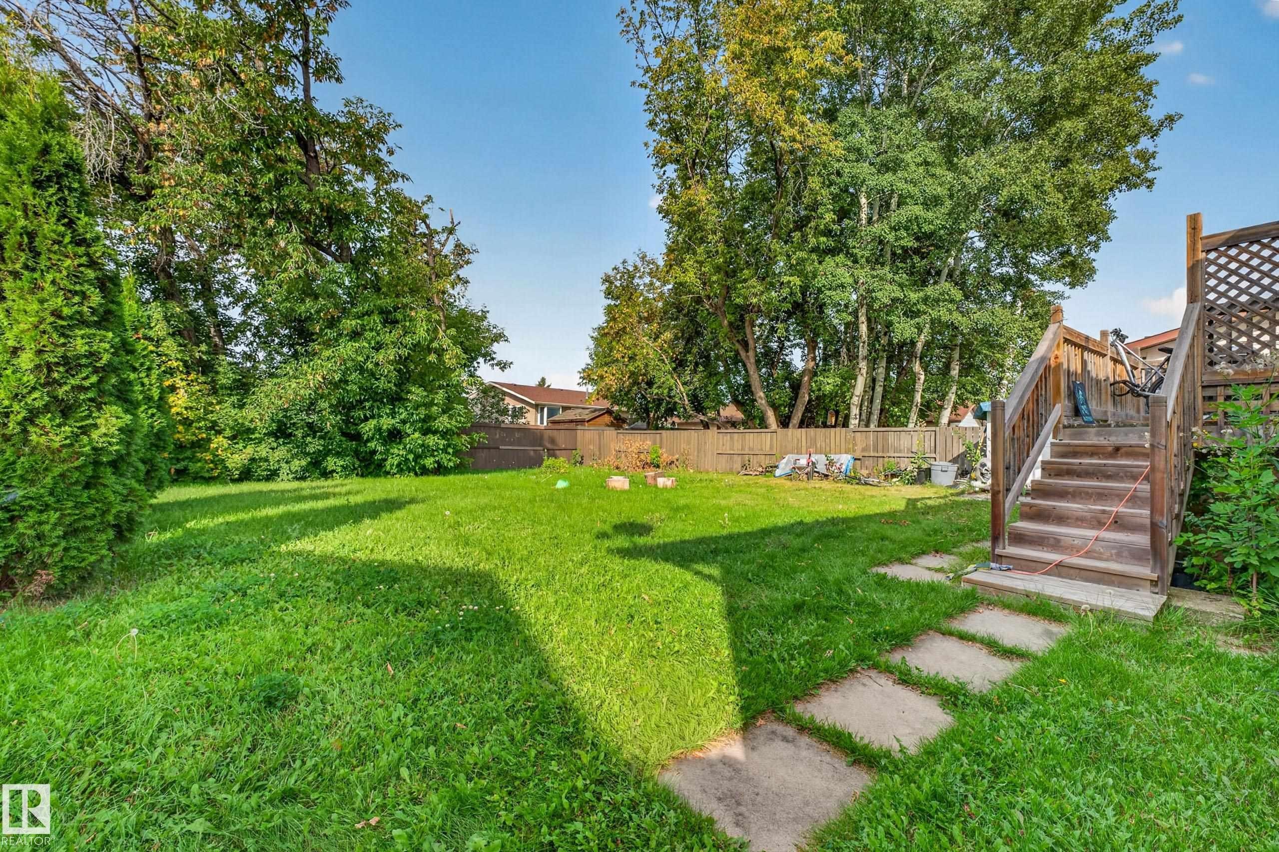 Expansive backyard featuring a lush lawn, mature trees, a wood-slat fence, and concrete stepping stones - 2412 80 Street Nw, Edmonton, AB
