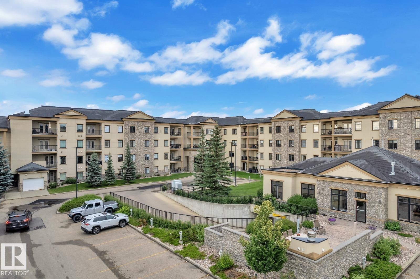 Exterior featuring stone and stucco finishes, multiple balconies, a paved parking area, a manicured lawn, and a stone patio - #105 160 Magrath Rd Nw, Edmonton, AB