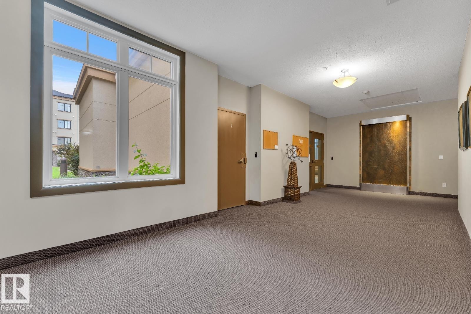 Spacious hallway featuring textured carpet flooring, a large multi-pane window, and a built-in wall water feature - #105 160 Magrath Rd Nw, Edmonton, AB