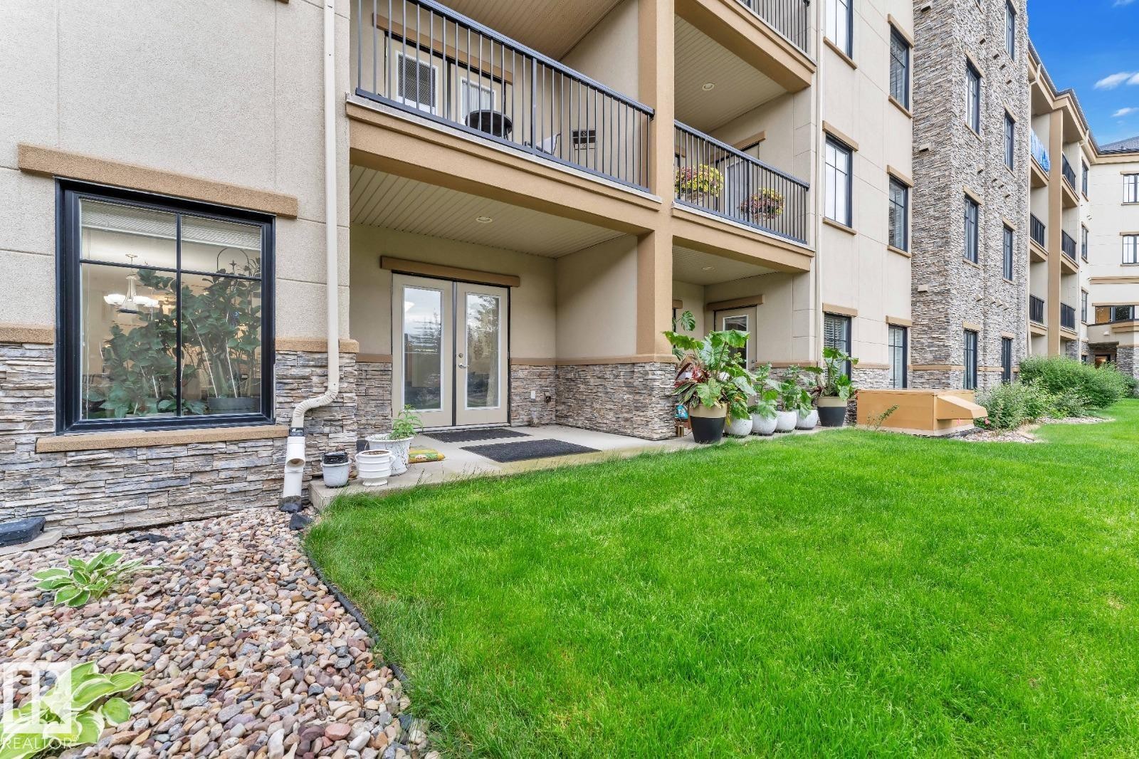 Ground-level patio with double glass doors, surrounded by stacked stone and stucco exterior - #105 160 Magrath Rd Nw, Edmonton, AB