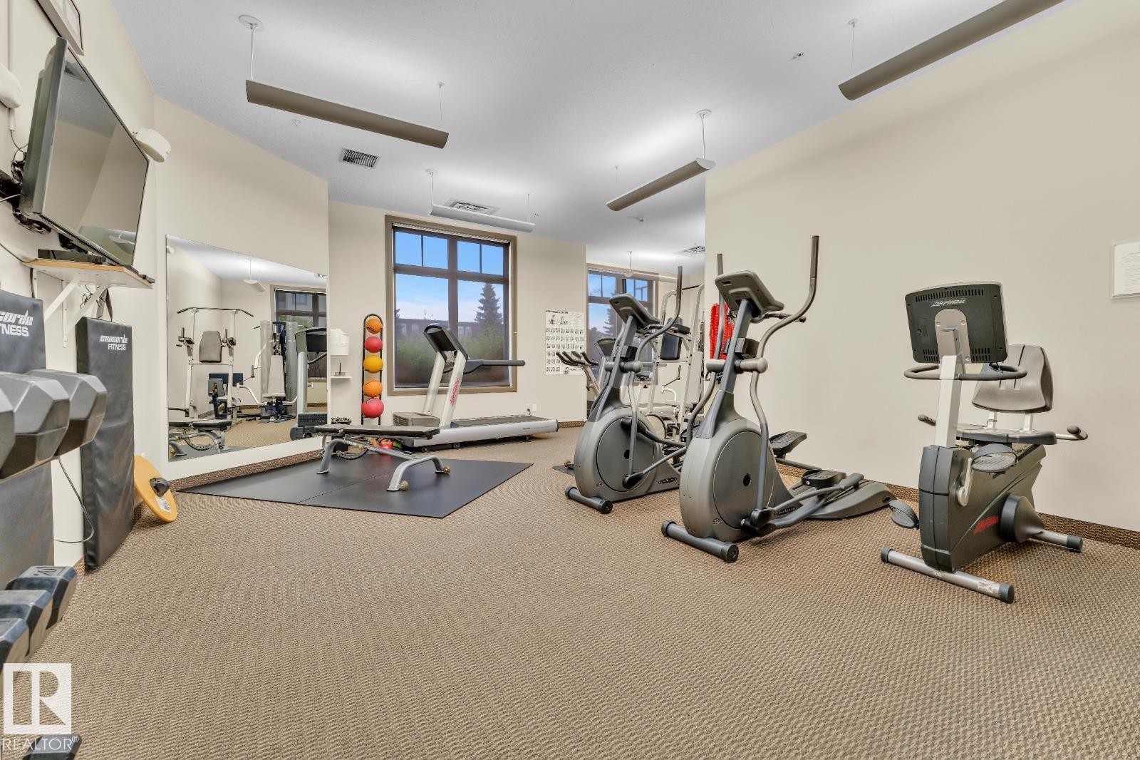 Carpeted fitness area with wall-mounted mirror, large window, and recessed lighting - #105 160 Magrath Rd Nw, Edmonton, AB