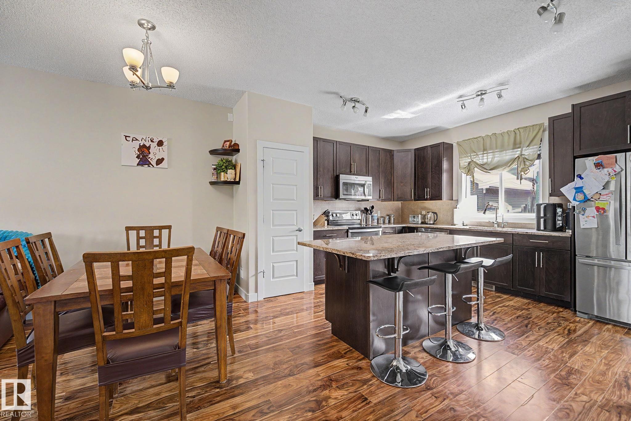 Open-concept kitchen and dining area featuring rich wood-finish flooring, dark wood cabinetry, a central island with granite countertop, and stainless steel appliances - 3343 Weidle Way Sw, Edmonton, AB