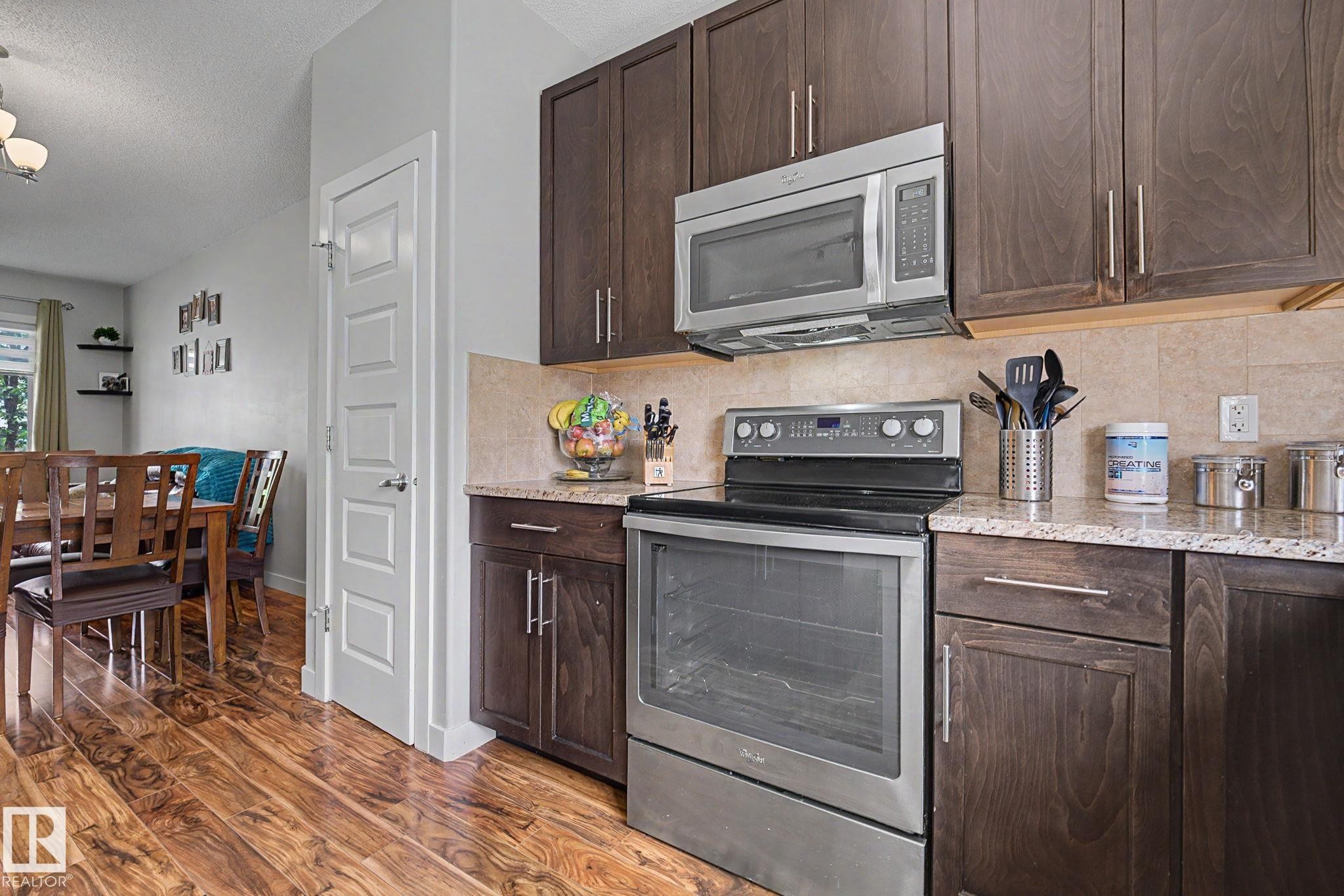 Kitchen featuring wood-finish flooring, dark wood cabinetry, light-toned granite countertops, a tiled backsplash, and stainless steel appliances - 3343 Weidle Way Sw, Edmonton, AB