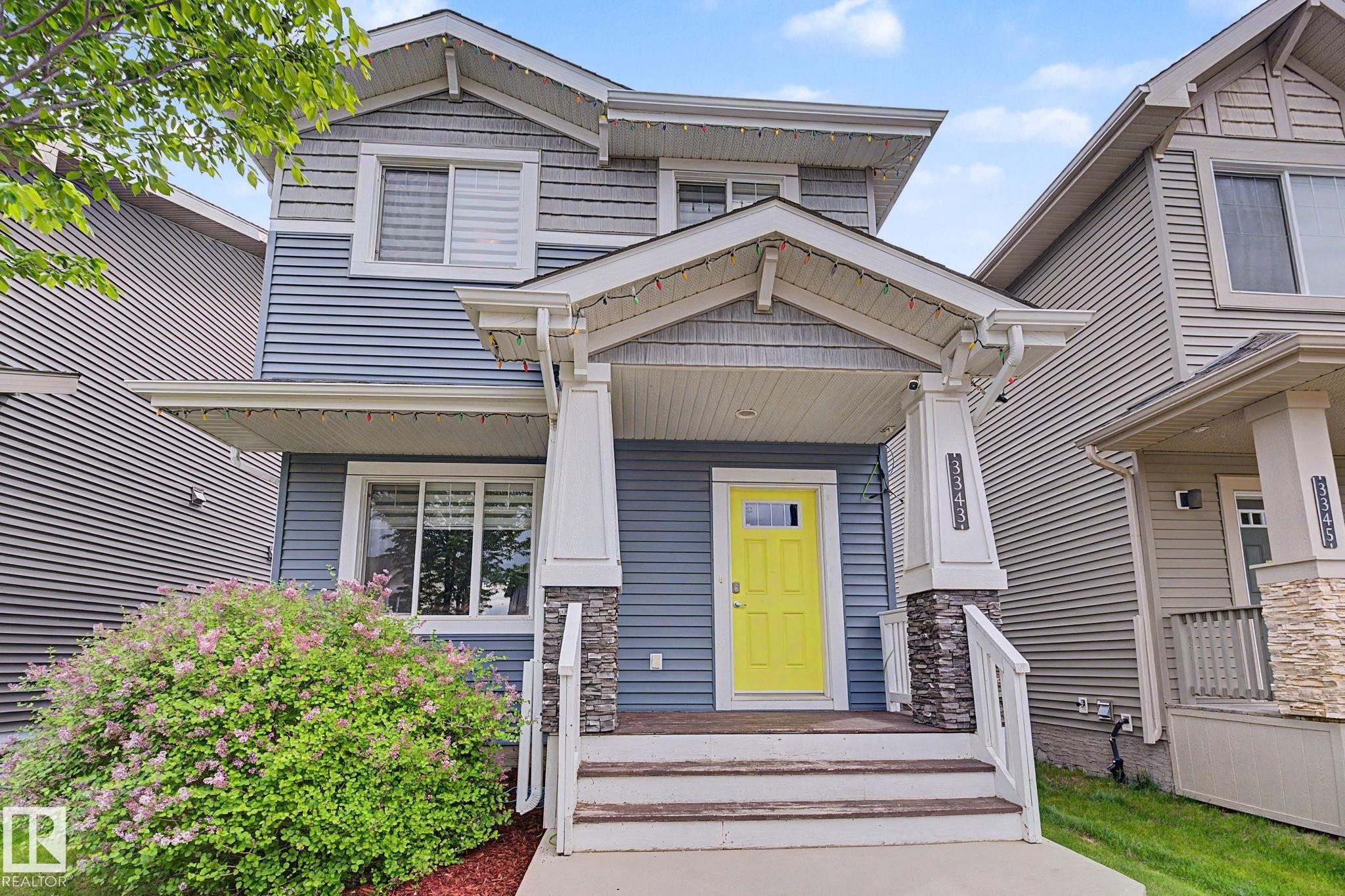 Front elevation featuring blue horizontal siding, decorative gray shake siding, a covered porch with stone-clad pillars, a bright yellow entry door, and a mature flowering shrub - 3343 Weidle Way Sw, Edmonton, AB