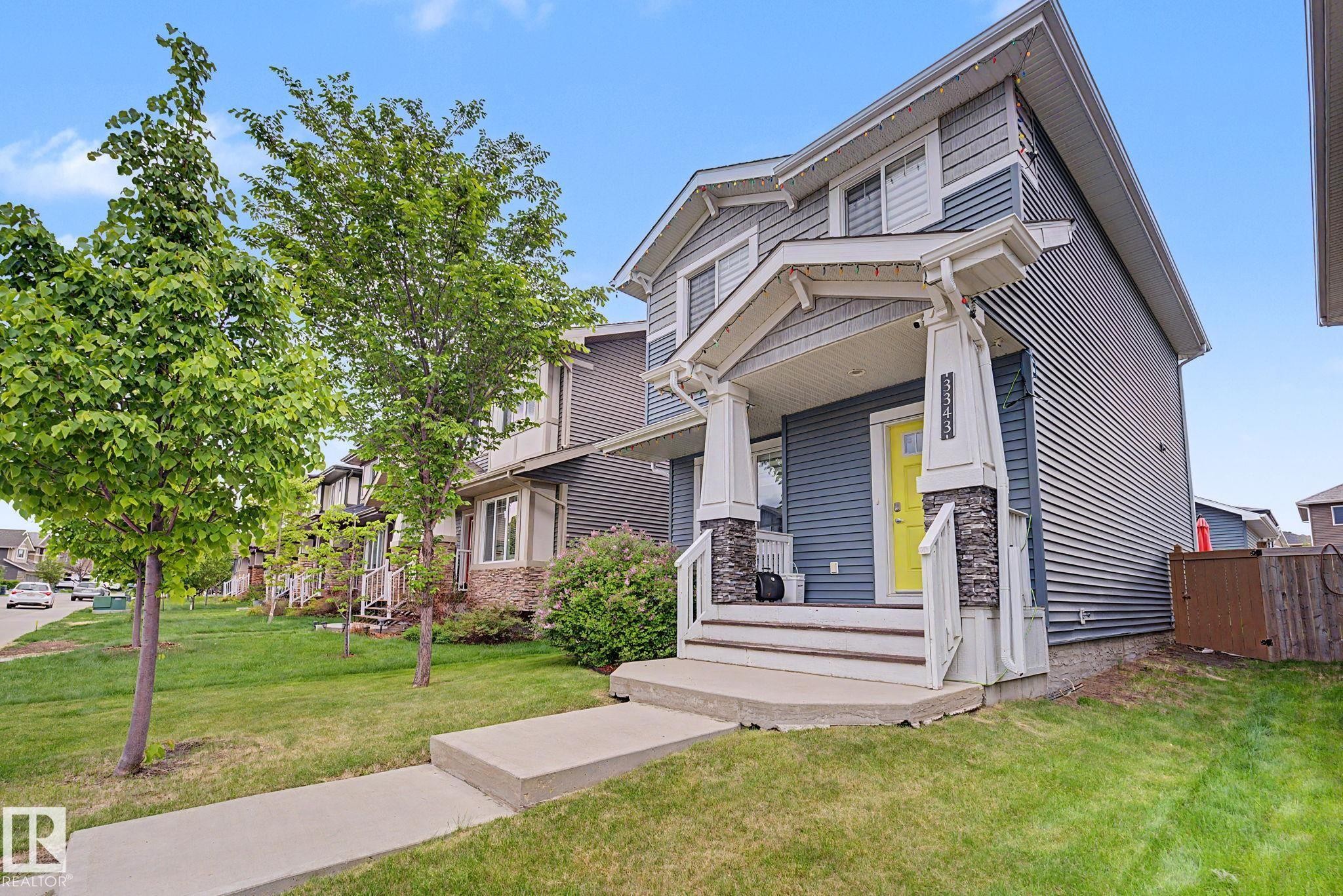 Two-story residence featuring a covered front porch with stone-clad pillars, a bright yellow entry door, and horizontal siding - 3343 Weidle Way Sw, Edmonton, AB