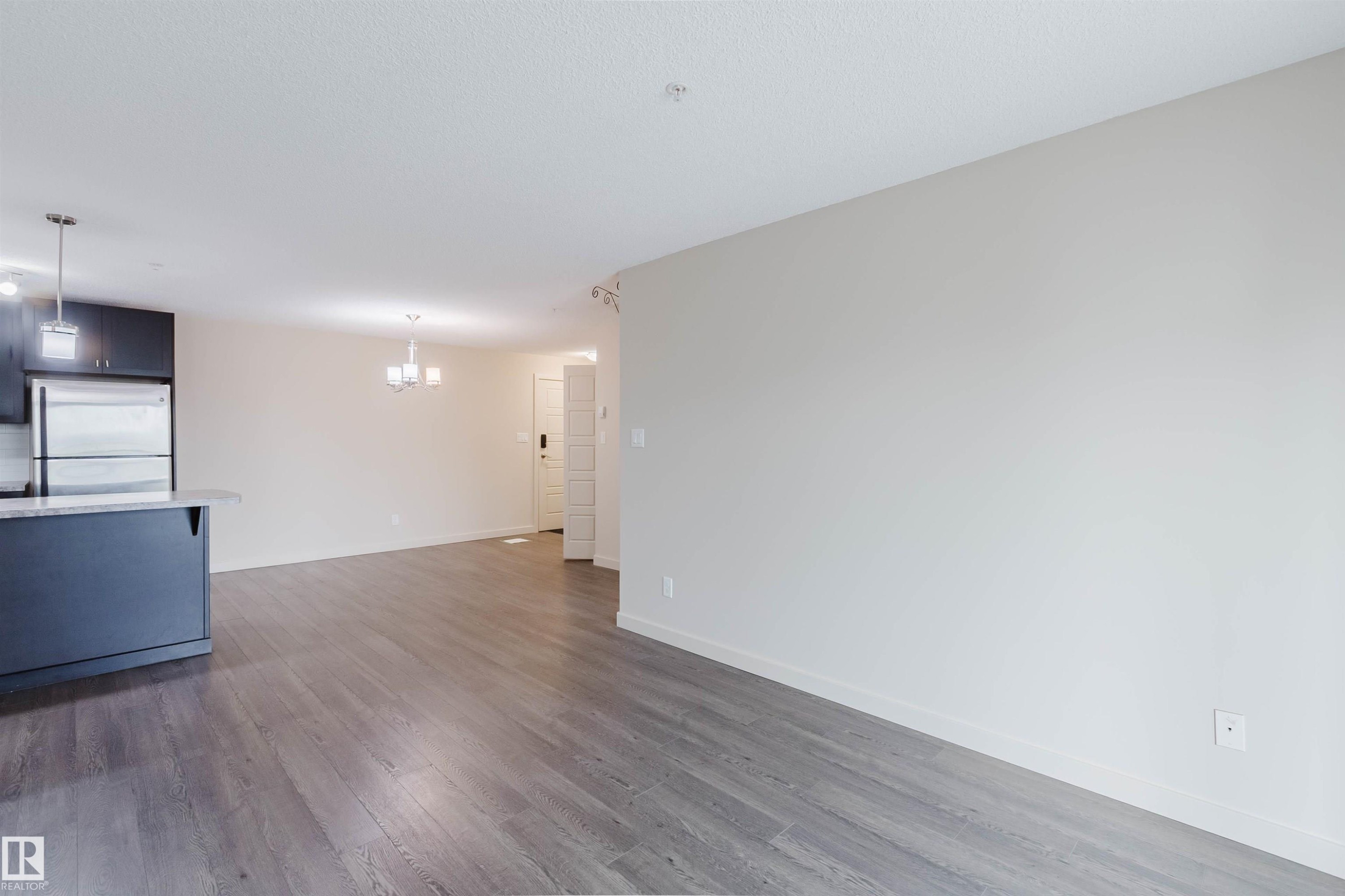 Expansive living area with wood-finish flooring, light neutral wall paint, and a modern chandelier - #304 6084 Stanton Dr Sw, Edmonton, AB - Indoor Photo Showing Kitchen