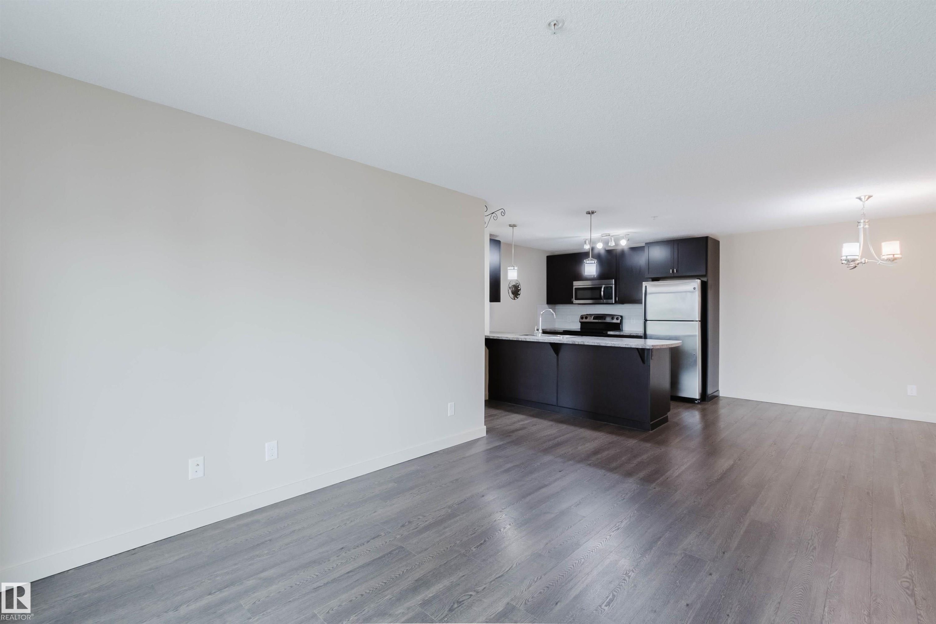 Open-concept living space featuring wood-finish flooring, a kitchen with dark cabinetry, stainless steel appliances, a breakfast bar, and pendant lighting - #304 6084 Stanton Dr Sw, Edmonton, AB - Indoor