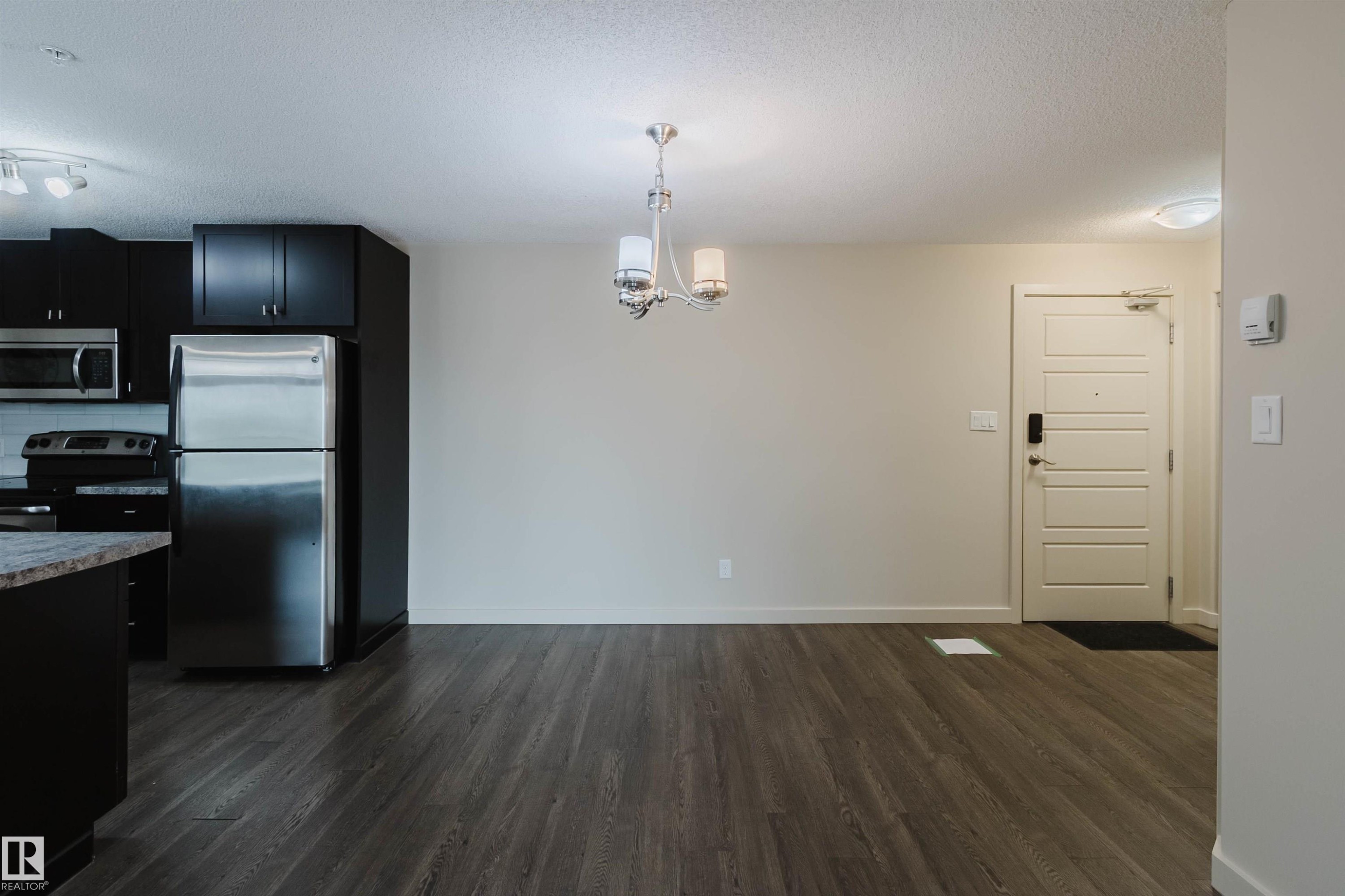 Open concept living space featuring dark wood-finish flooring, a contemporary chandelier, a white paneled entrance door, and a modern kitchen with black cabinetry and stainless steel appliances - #304 6084 Stanton Dr Sw, Edmonton, AB - Indoor Photo Showing Kitchen