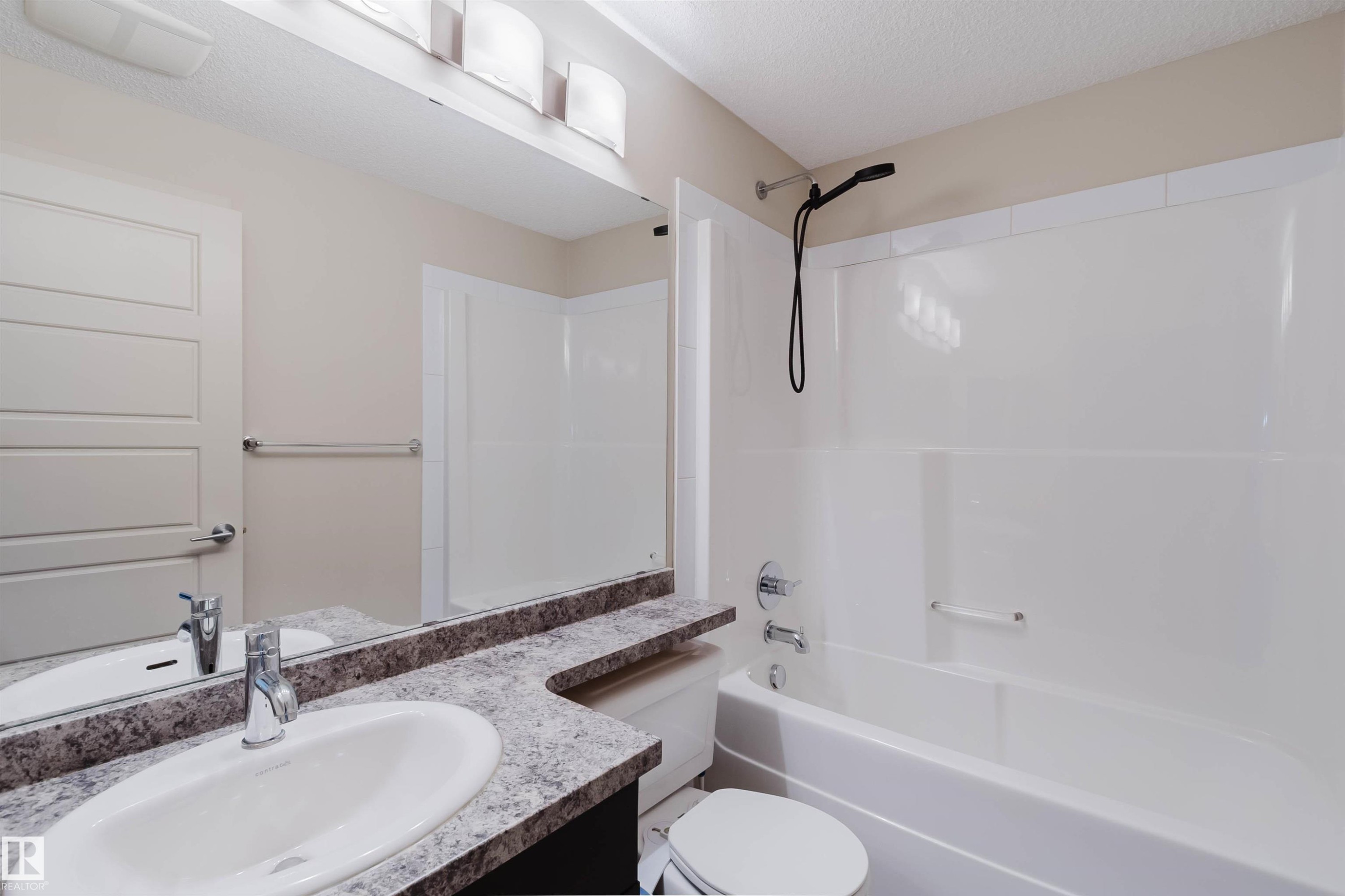 Bathroom featuring a white drop-in sink with a chrome faucet, a speckled countertop, an oversized wall mirror, and a full bathtub with an integrated shower and black showerhead - #304 6084 Stanton Dr Sw, Edmonton, AB - Indoor Photo Showing Bathroom