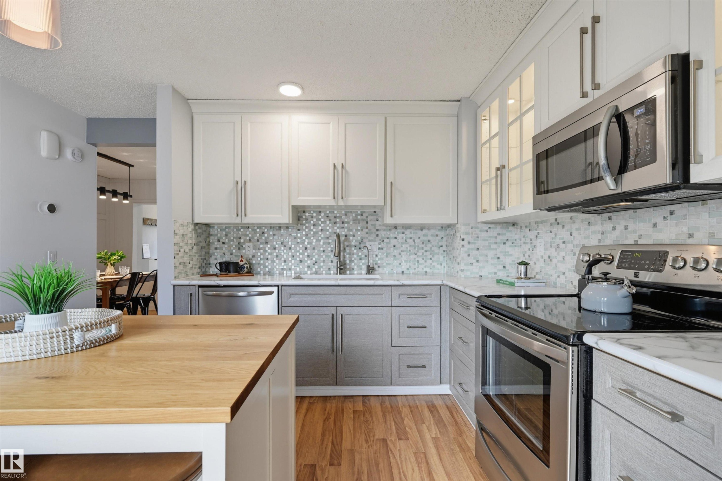 Modern kitchen featuring stainless steel appliances, mosaic tile backsplash, wood-finish flooring, white upper cabinetry, and gray lower cabinetry - 4125 38 Street Nw, Edmonton, AB