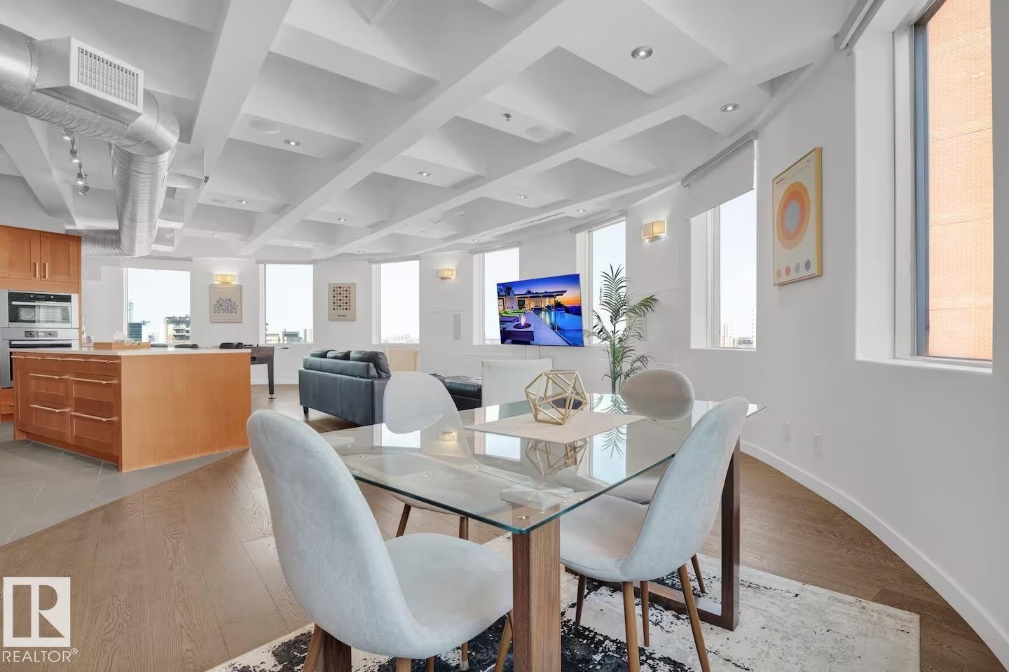 Open-concept living space featuring wood-finish flooring, a coffered ceiling, exposed ductwork, a kitchen island with integrated appliances, and multiple windows - #701 10105 109 St Nw, Edmonton, AB