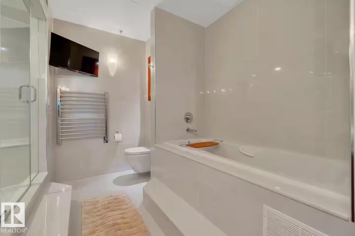 Built-in tub with chrome fixtures, glass-enclosed shower, wall-mounted television, chrome towel warmer, and wall-mounted toilet - #701 10105 109 St Nw, Edmonton, AB
