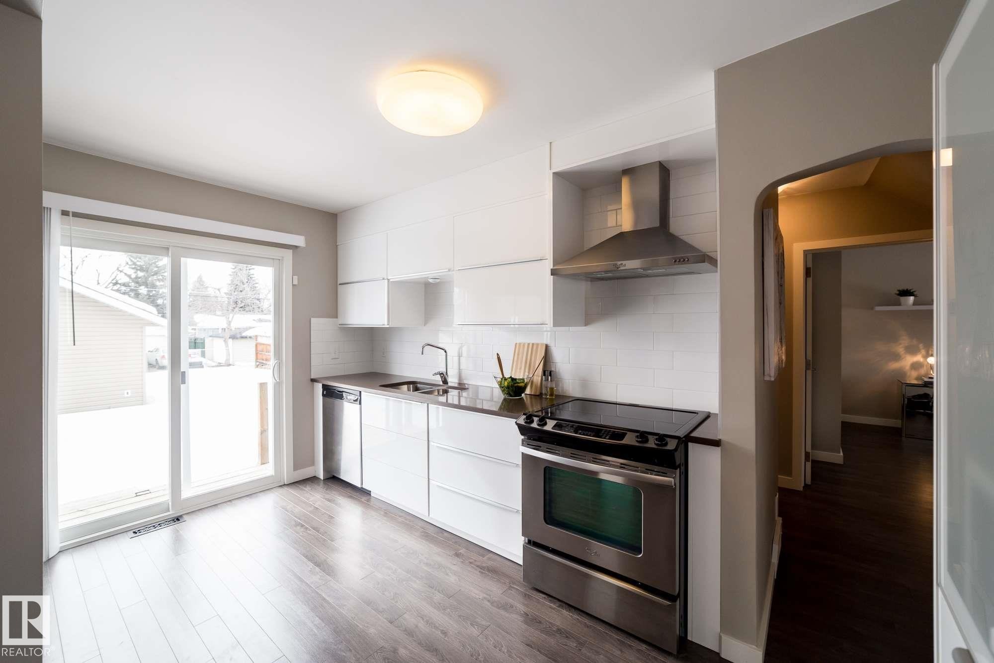 Modern kitchen with wood-finish flooring, white cabinetry, stainless steel appliances, and a sliding glass door - 12228 134 Street Nw, Edmonton, AB