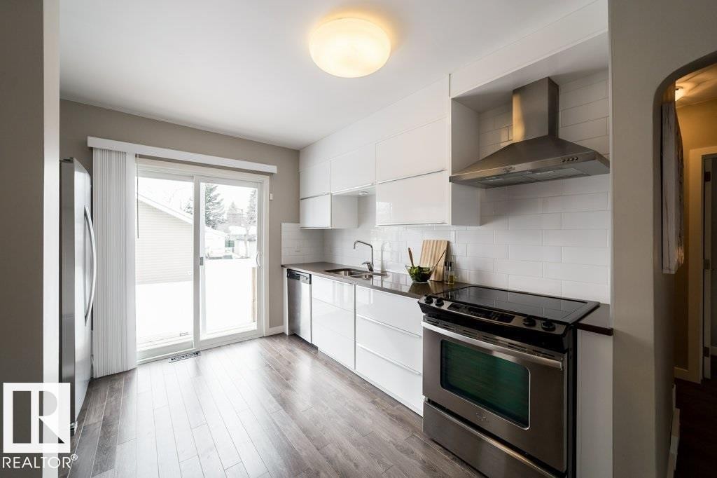 Kitchen featuring stainless steel appliances, white flat-panel cabinetry, dark countertops, white subway tile backsplash, and wood-finish flooring - 12228 134 Street Nw, Edmonton, AB