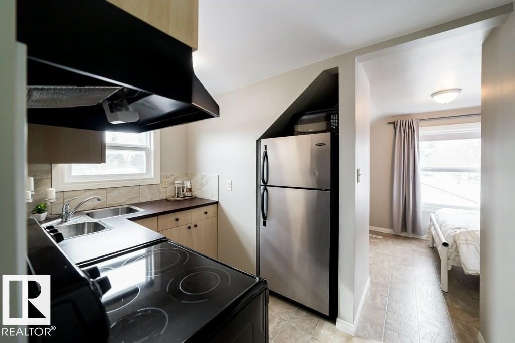 Compact kitchen area featuring a built-in stainless steel refrigerator, black electric stovetop, double basin stainless steel sink, and light wood-finish cabinetry - 12228 134 Street Nw, Edmonton, AB