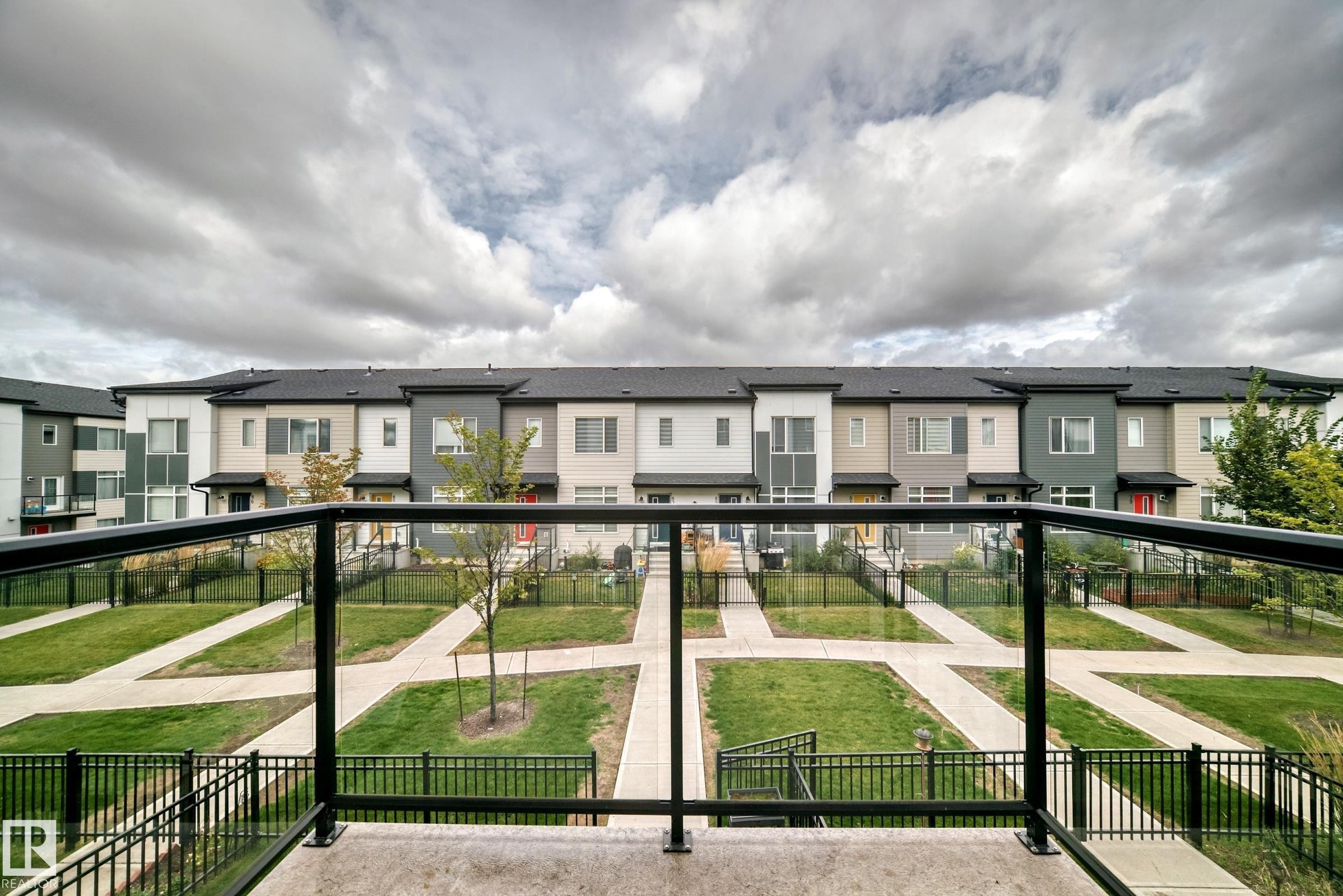 Black metal and glass panel railing with a concrete deck, overlooking a structured green space with walkways and multi-story residential buildings featuring various exterior finishes - #56 804 Welsh Dr Sw, Edmonton, AB