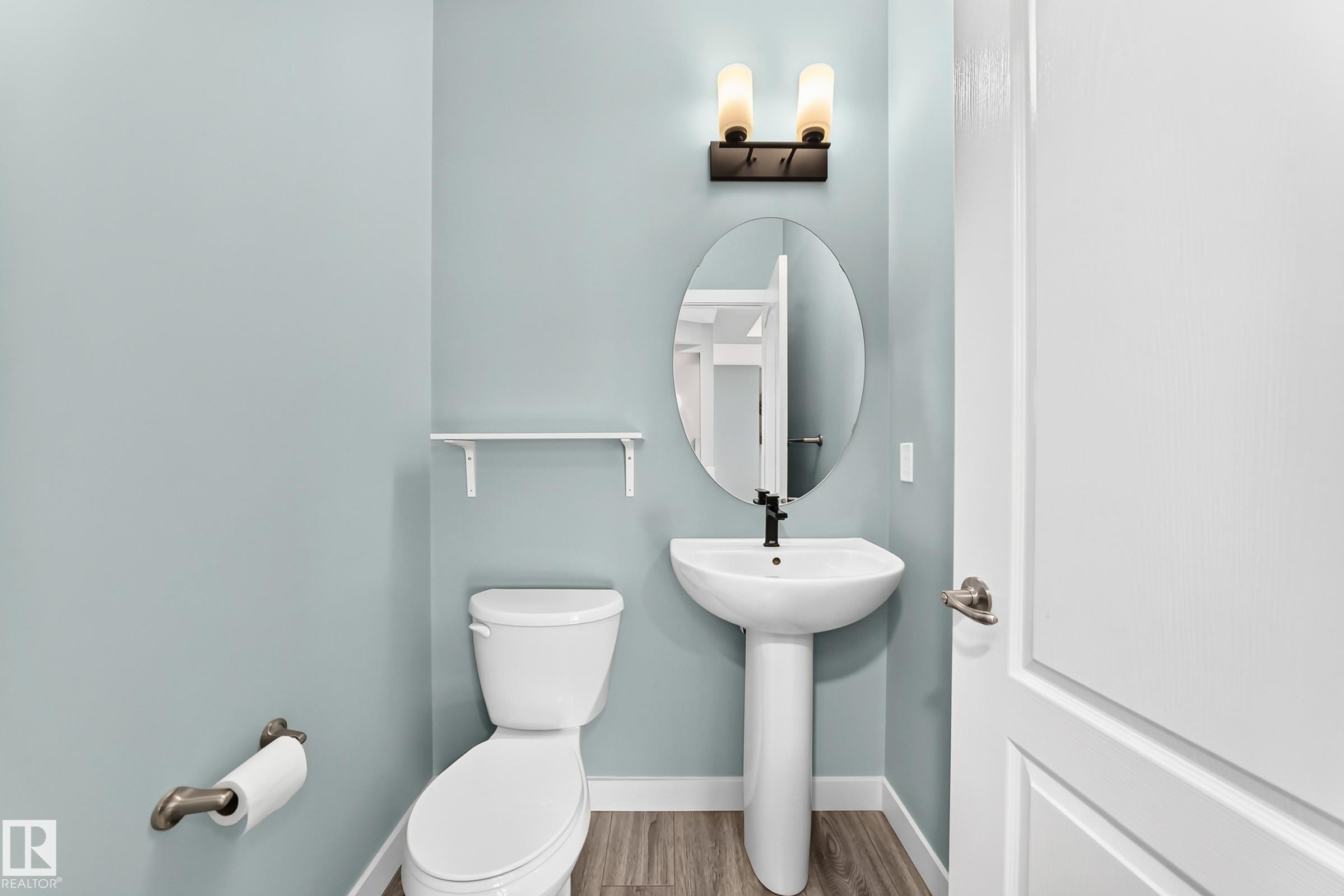 Pedestal sink with a matte black faucet, oval mirror, wall-mounted sconce lighting, and wood-finish flooring - #56 804 Welsh Dr Sw, Edmonton, AB