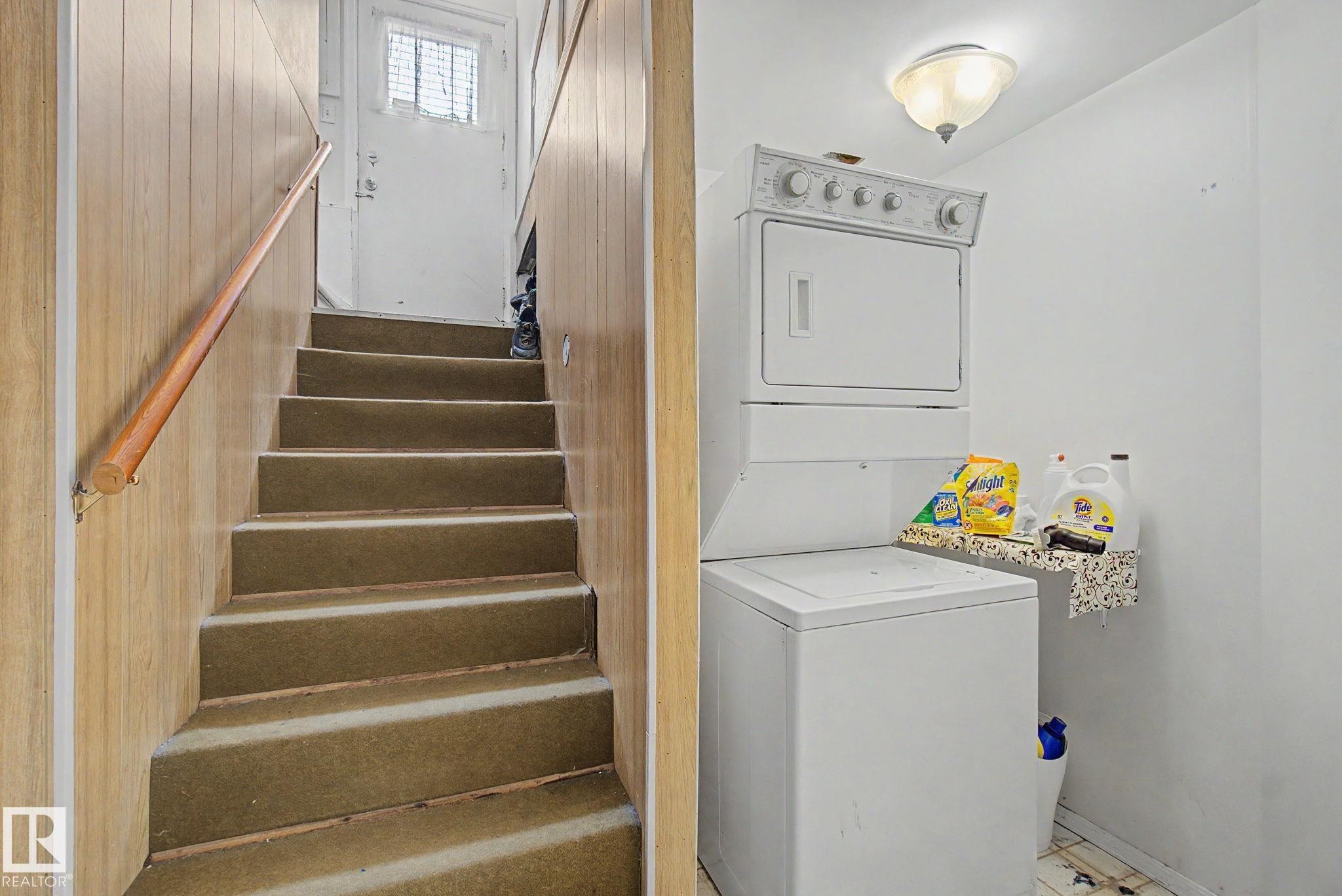 Dedicated laundry area featuring a stacked washer and dryer, adjacent to a carpeted staircase with a wooden handrail and paneled walls - 8710 83 Avenue Nw, Edmonton, AB - Indoor Photo Showing Laundry Room