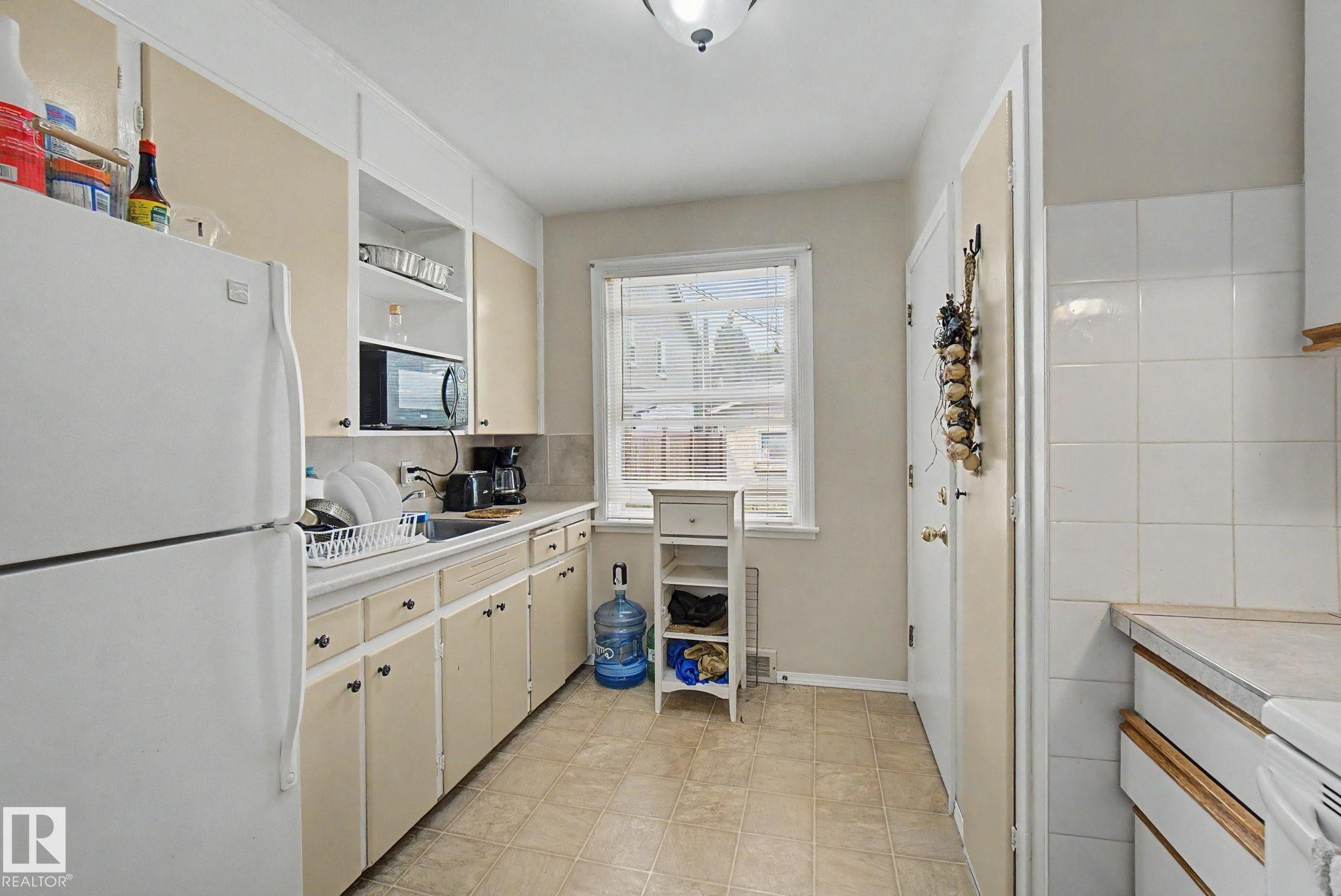 Galley kitchen featuring light-toned cabinetry, a window with blinds, and vinyl flooring - 8710 83 Avenue Nw, Edmonton, AB - Indoor Photo Showing Kitchen