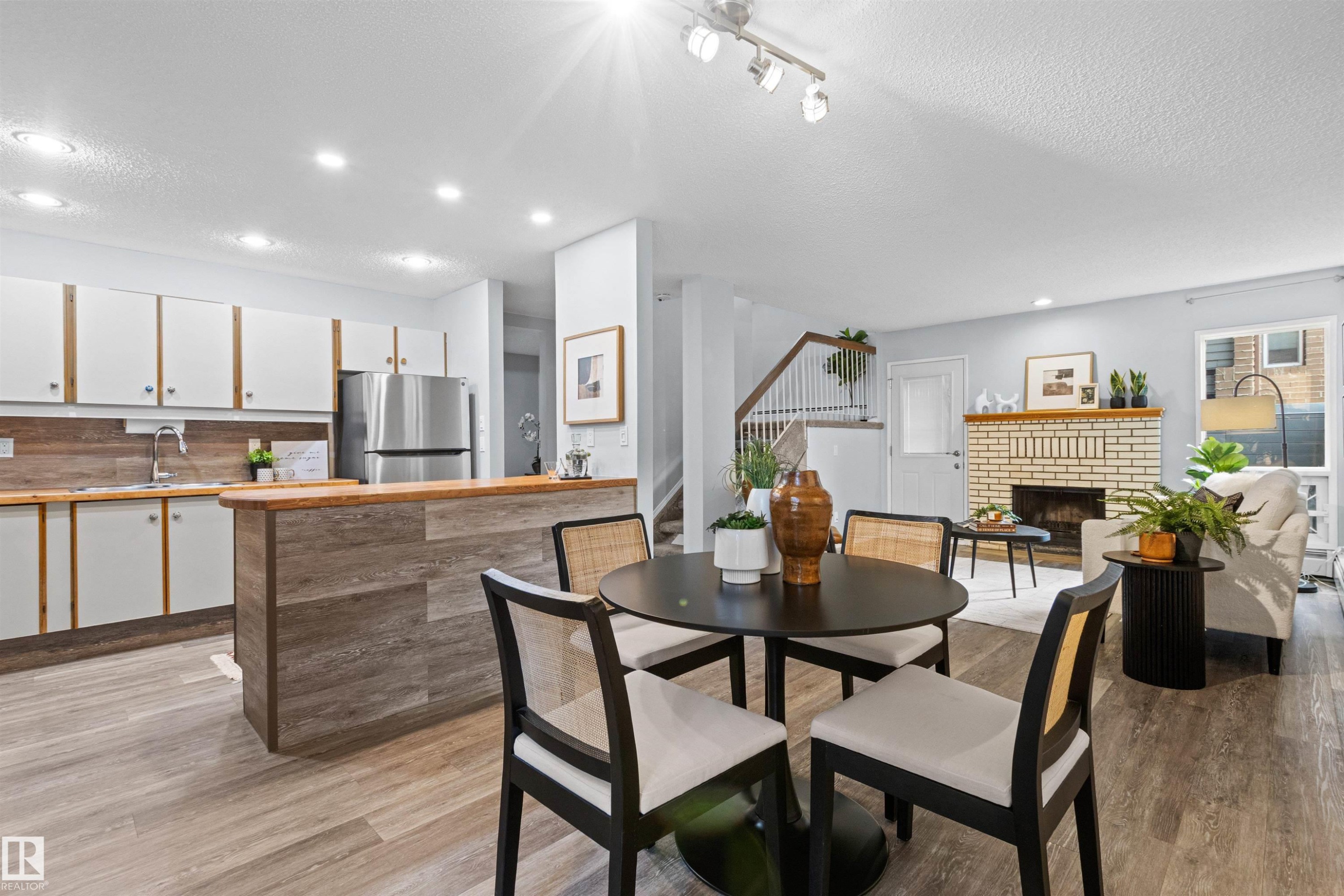 Open-concept living area featuring wood-finish flooring, a kitchen with stainless steel appliances, a brick fireplace, and track lighting - #7 10032 113 St Nw, Edmonton, AB