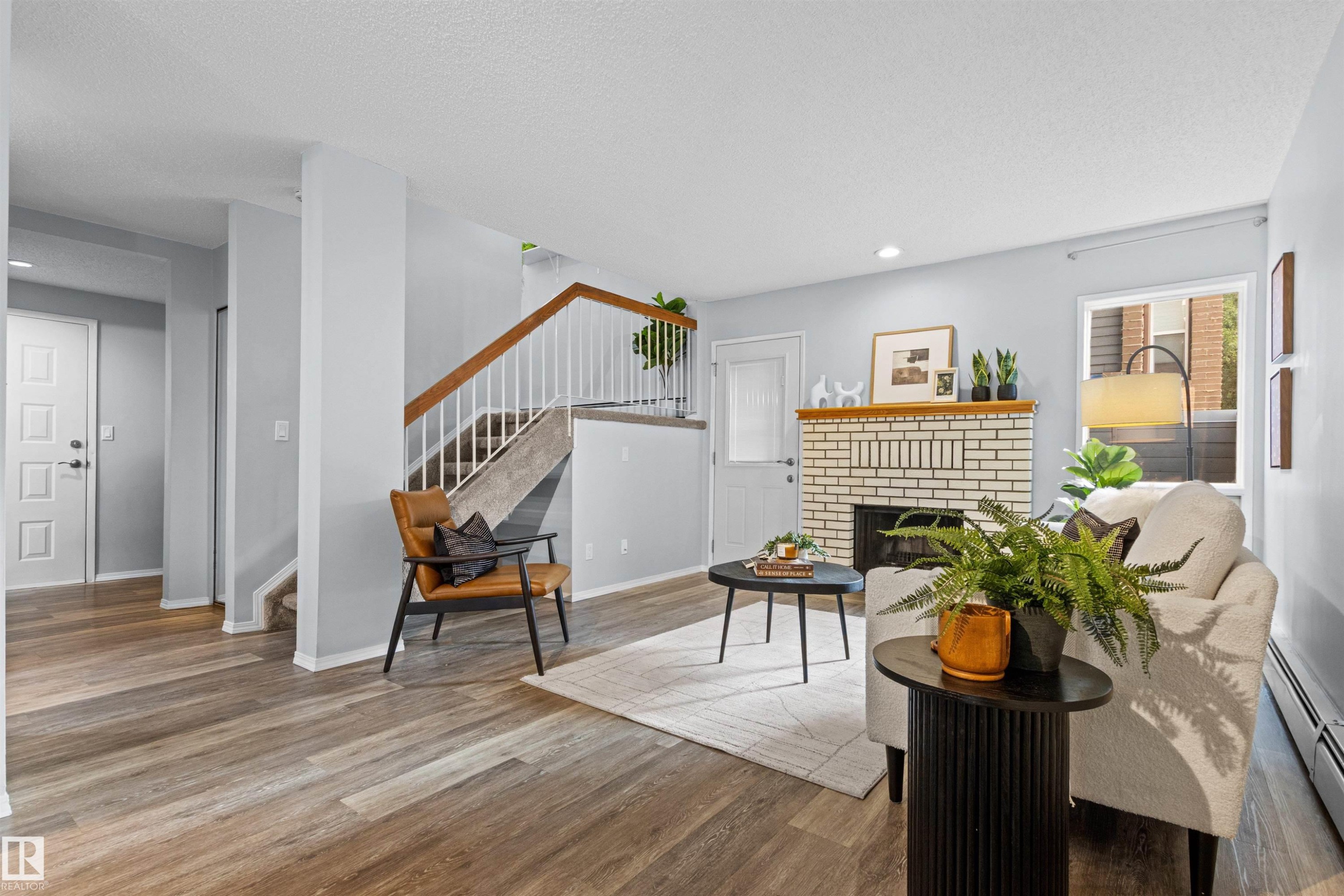 Wood-finish flooring throughout, featuring a white brick fireplace with a wood mantle, a staircase with white spindles and wood handrail, and white panel doors - #7 10032 113 St Nw, Edmonton, AB