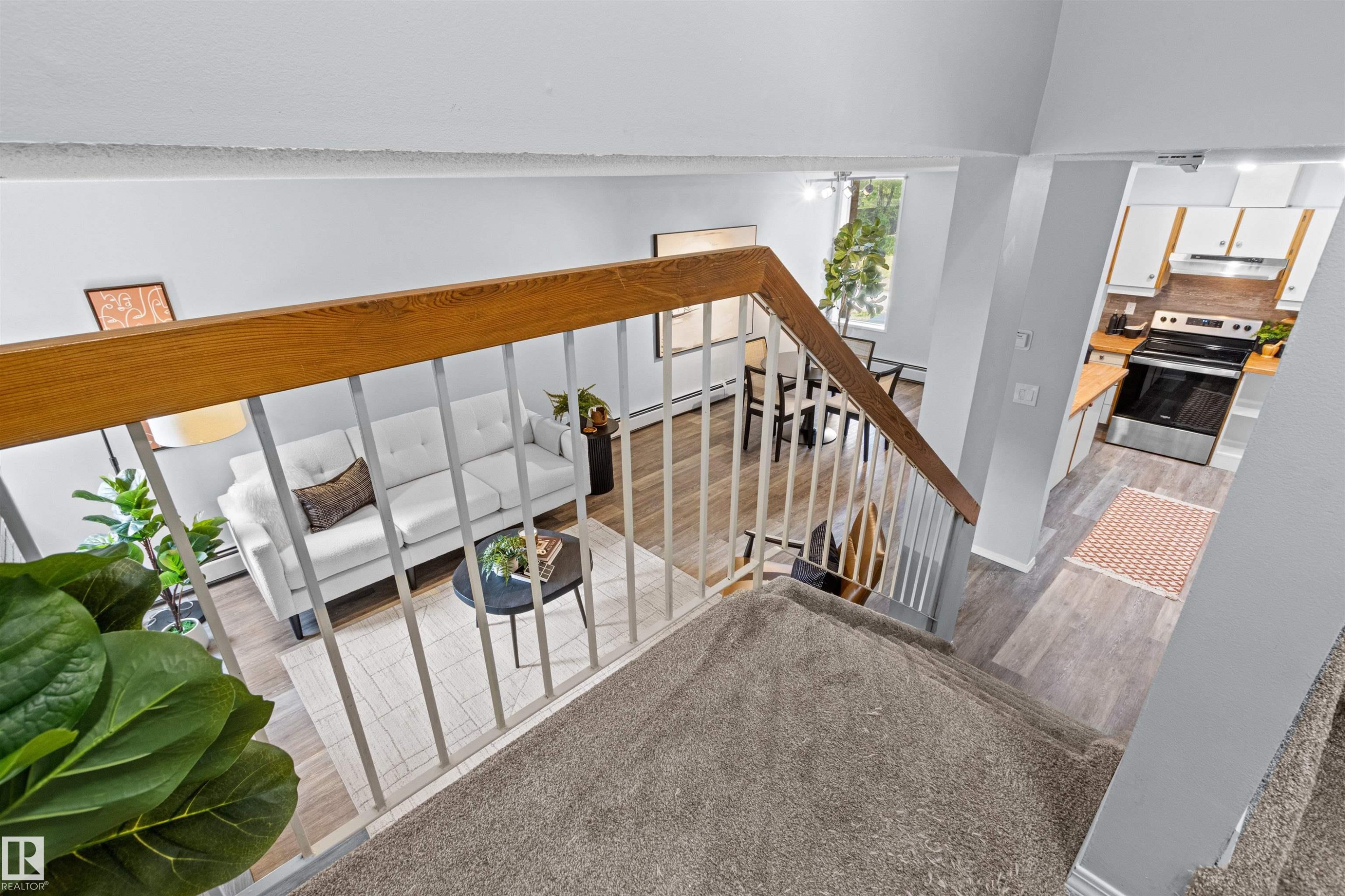Carpeted staircase with wood railing, leading to an open-concept living area with wood-finish flooring, a dining space, and a kitchen featuring white cabinetry, light countertops, and stainless steel appliances - #7 10032 113 St Nw, Edmonton, AB