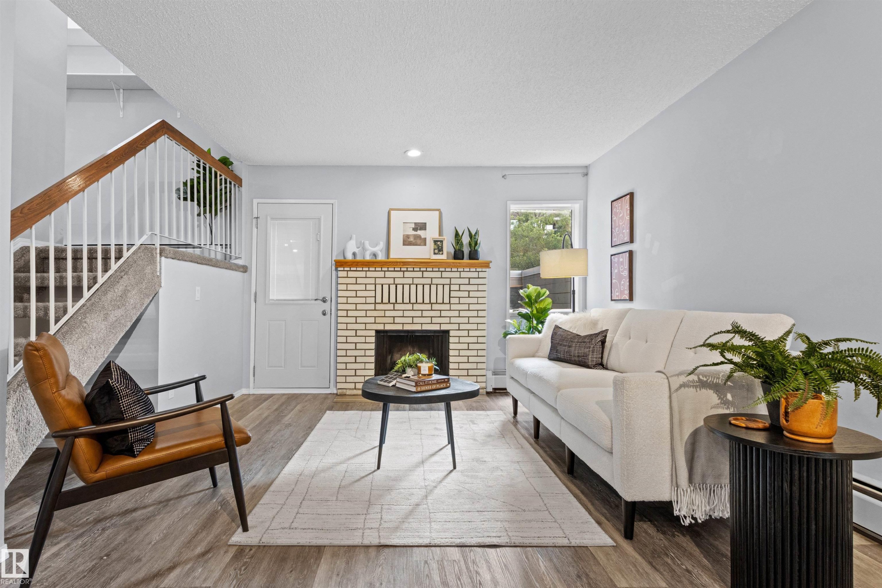 Light-colored brick fireplace with hearth, wood-finish flooring, staircase with carpeted treads and wood handrail, and white paneled door with glass insert - #7 10032 113 St Nw, Edmonton, AB