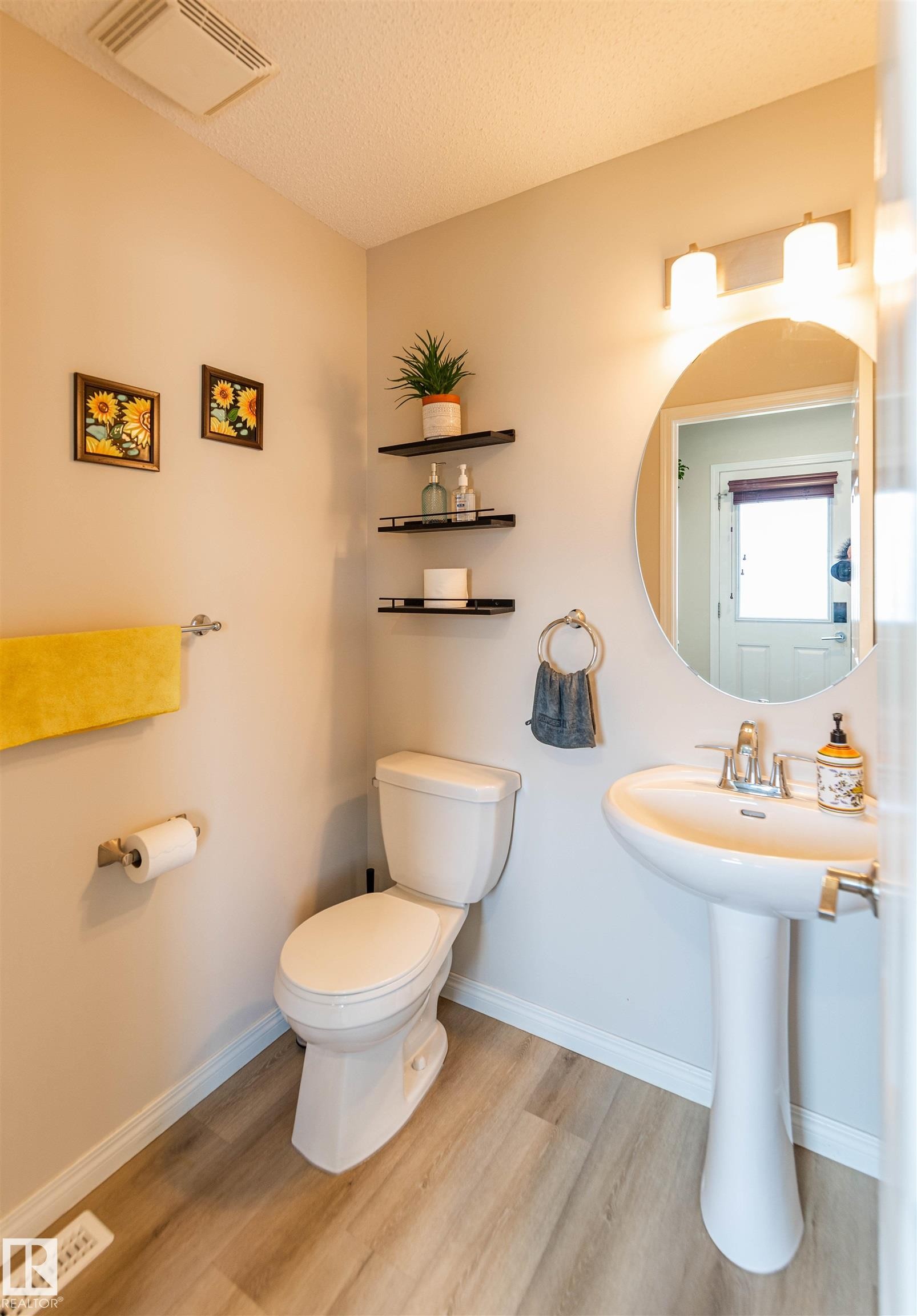 Pedestal sink with chrome fixtures, wood-finish flooring, wall-mounted oval mirror, and three-light vanity bar - 8527 Ellis Link Nw, Edmonton, AB - Indoor Photo Showing Bathroom