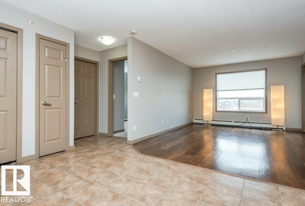 Expansive room featuring wood-finish flooring, a large window with roller shade, and neutral wall tones - 401 5005 165 Avenue Nw, Edmonton, AB