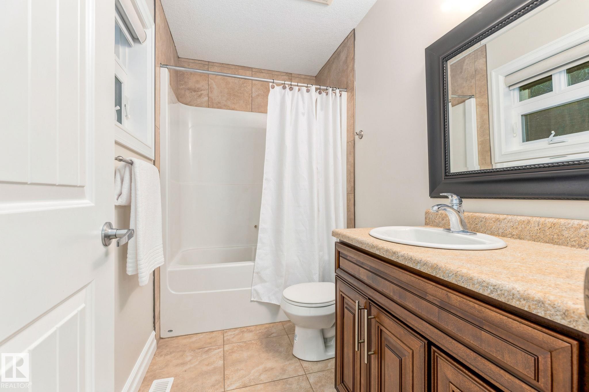 Bathroom featuring a warm-toned tile floor, a shower-tub combination with tile surround, a dark wood-finish vanity with an undermount sink, and a framed mirror - 10143 123 Street Nw, Edmonton, AB
