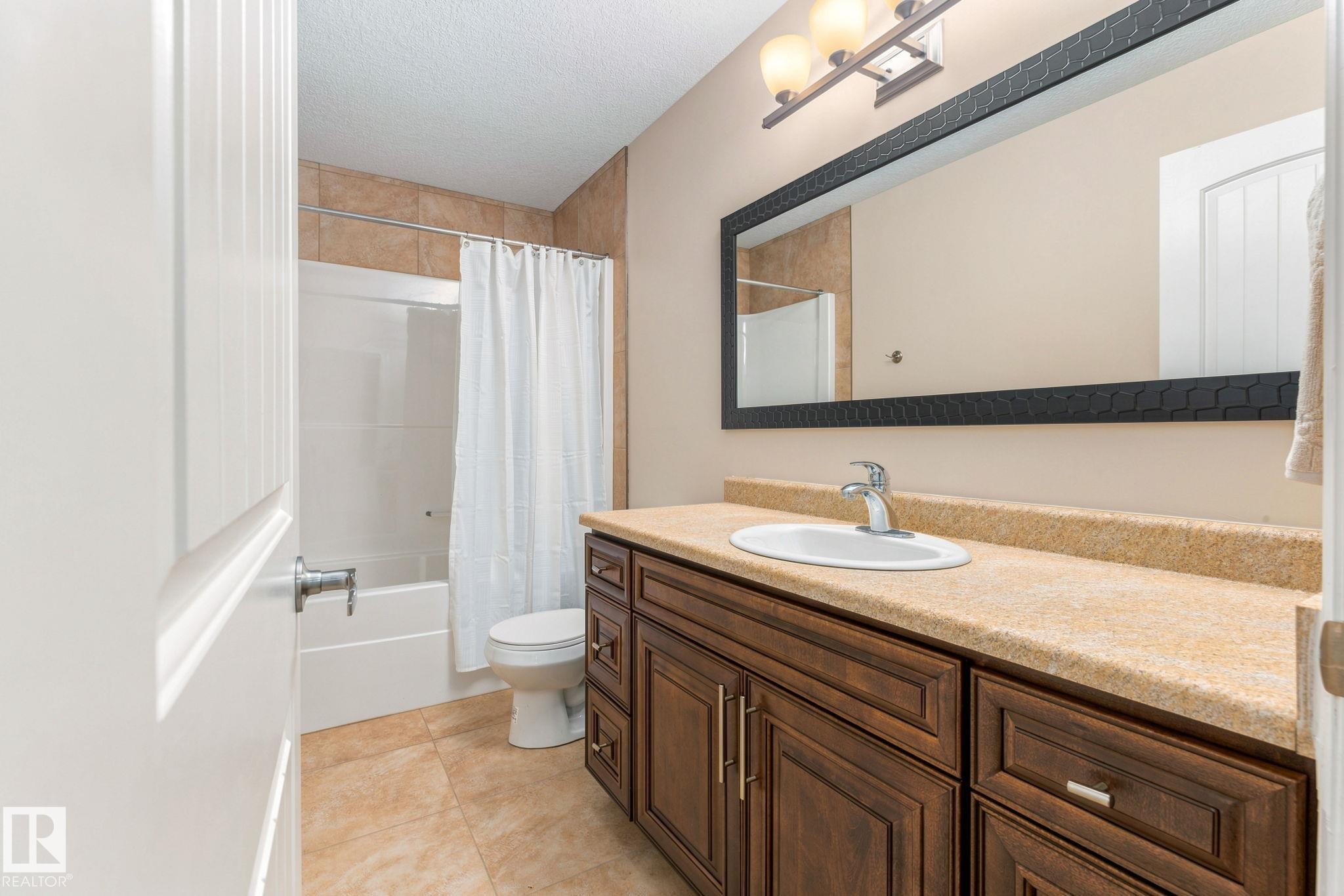 Bathroom featuring a wood-finish vanity with an integrated sink, a large framed mirror, a three-light vanity fixture, a tiled shower-tub combination, and tiled flooring - 10143 123 Street Nw, Edmonton, AB