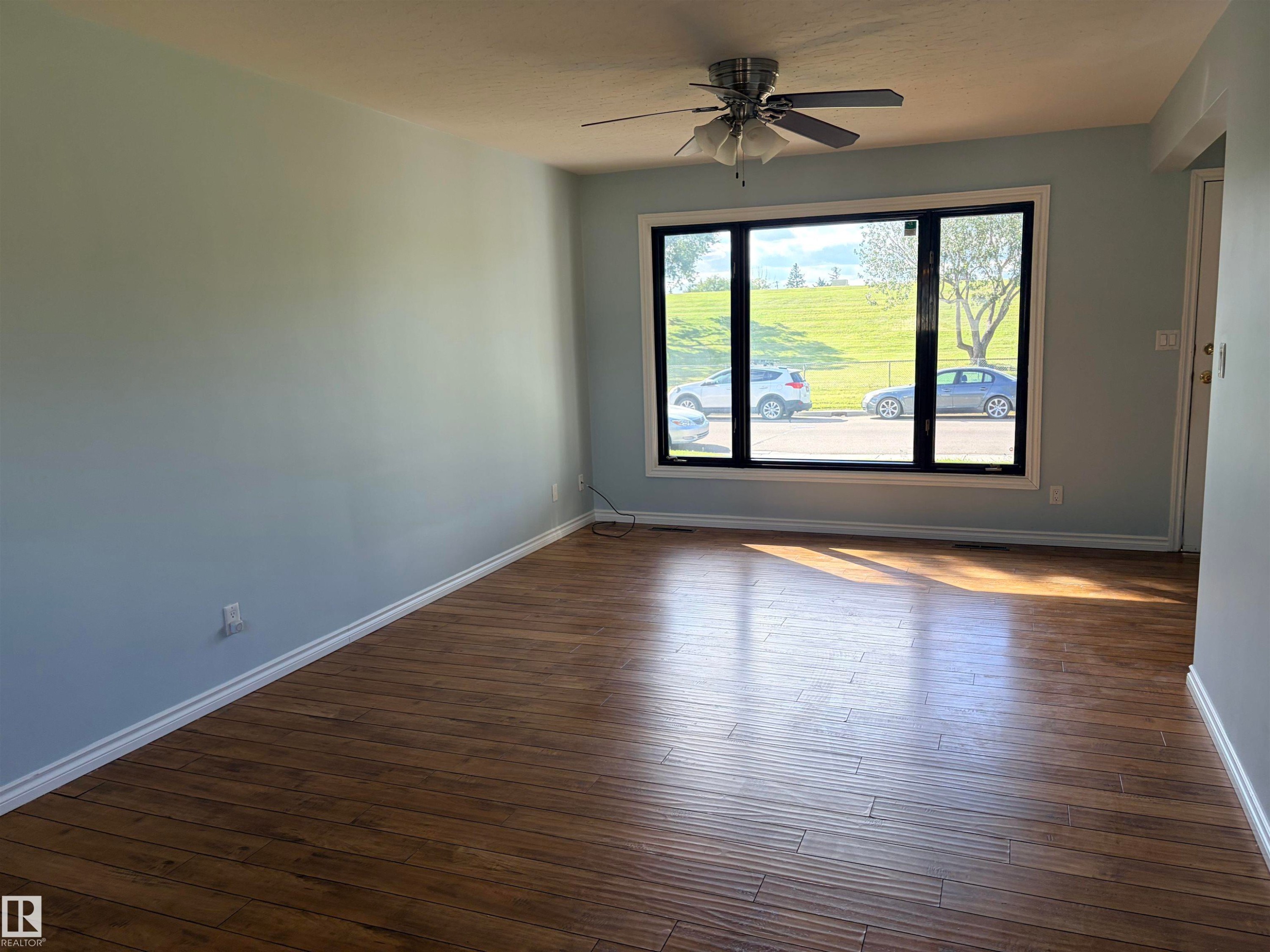 Wood-finish flooring, light blue wall paint, a ceiling fan, and a large window with black framing Main living area . - 13219 107 Street Nw, Edmonton, AB