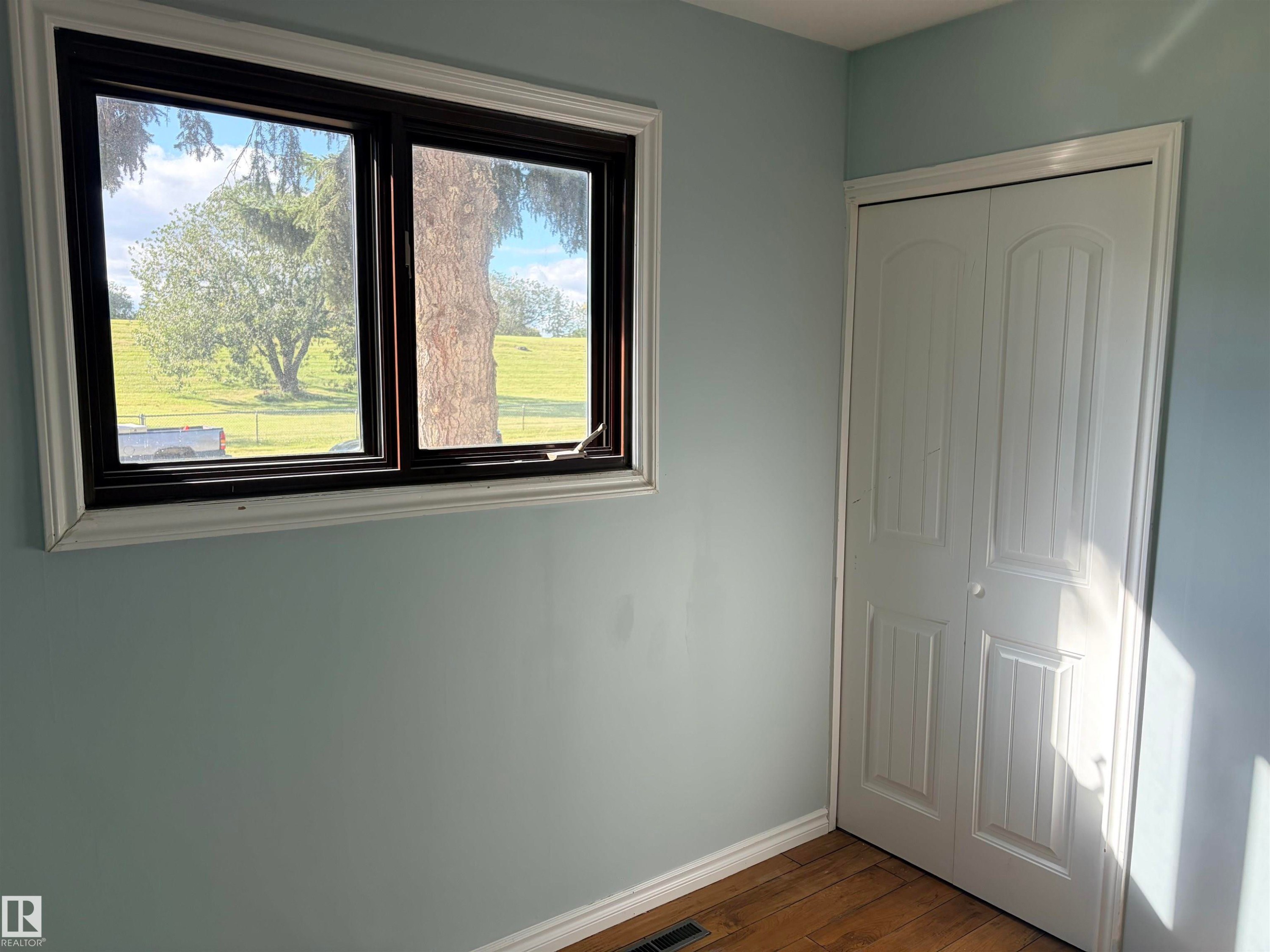 Wood-finish flooring, light blue wall paint, white bi-fold closet doors, and dark-framed windows with white trim - 13219 107 Street Nw, Edmonton, AB
