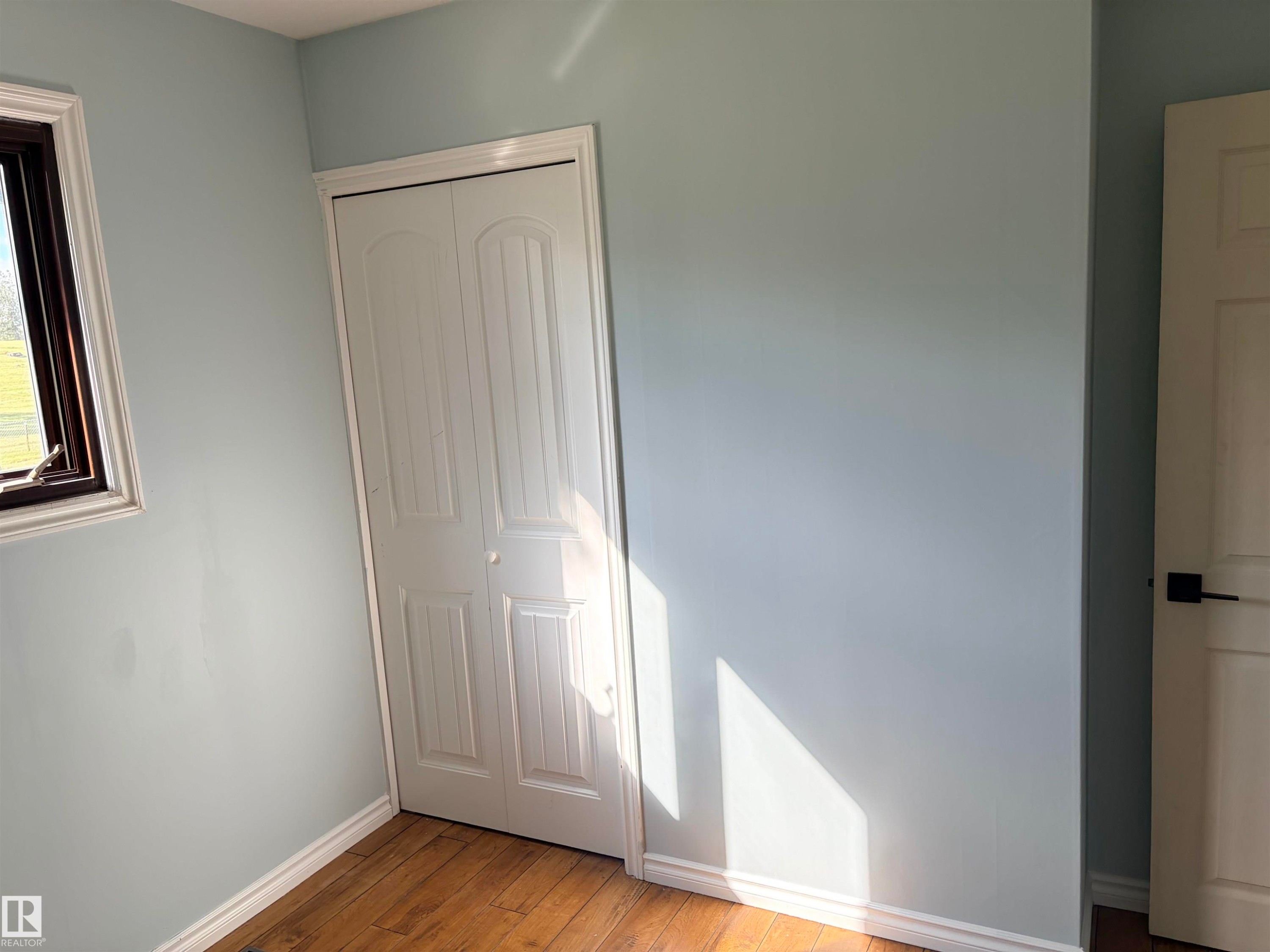 Wood-finish flooring, light blue wall paint, white trim, dark framed window, and white bi-fold closet doors Bedroom 2 - 13219 107 Street Nw, Edmonton, AB