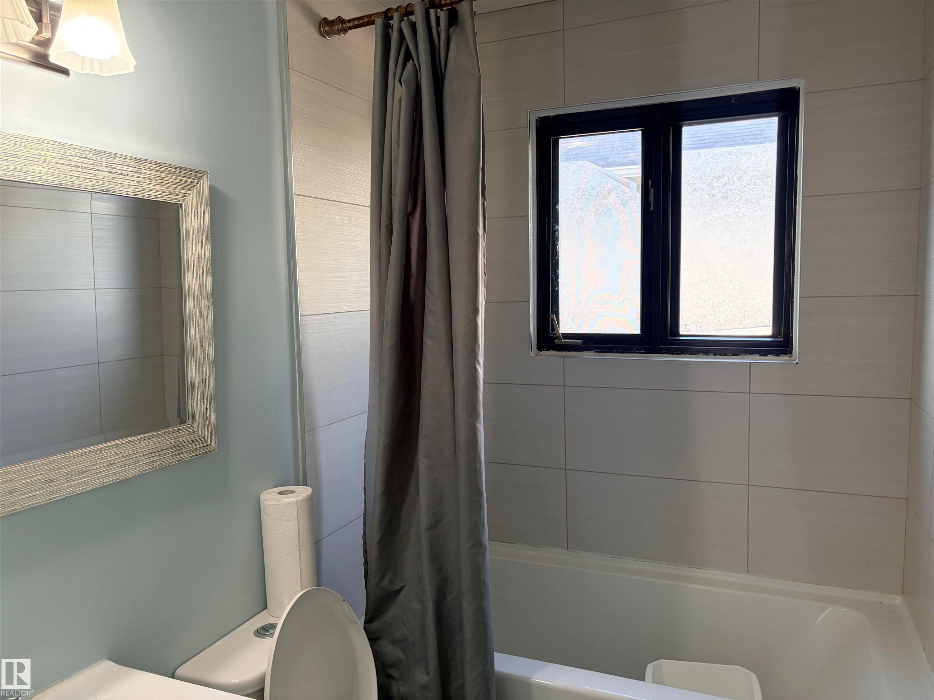 Bathtub with tiled surround, a dark-framed window, and a wall-mounted light fixture Upstairs main bath - 13219 107 Street Nw, Edmonton, AB