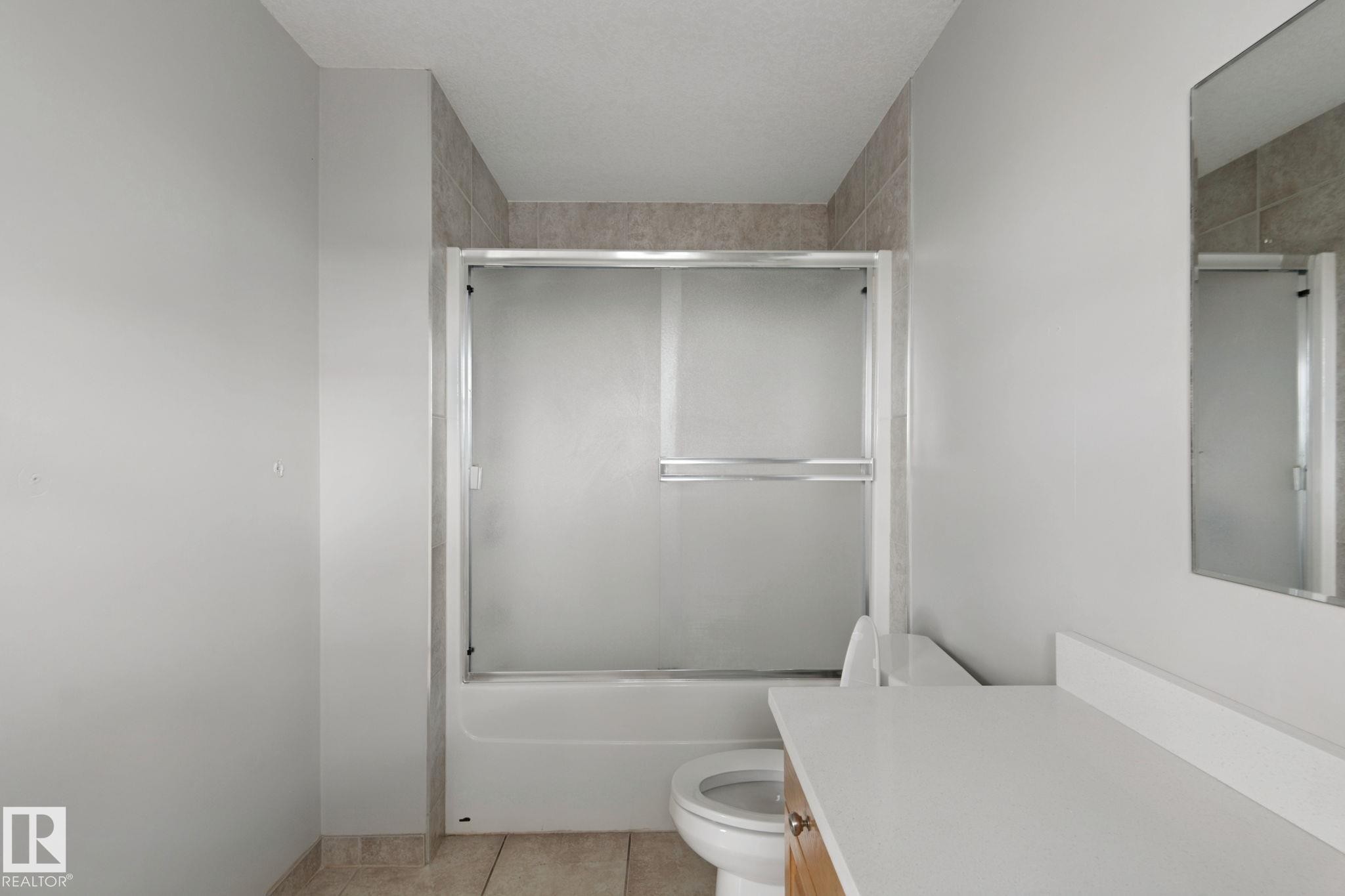 Bathtub with sliding frosted glass enclosure, tiled shower surround, light-toned countertop, and reflective wall mirror - 16807 78 Street Nw, Edmonton, AB