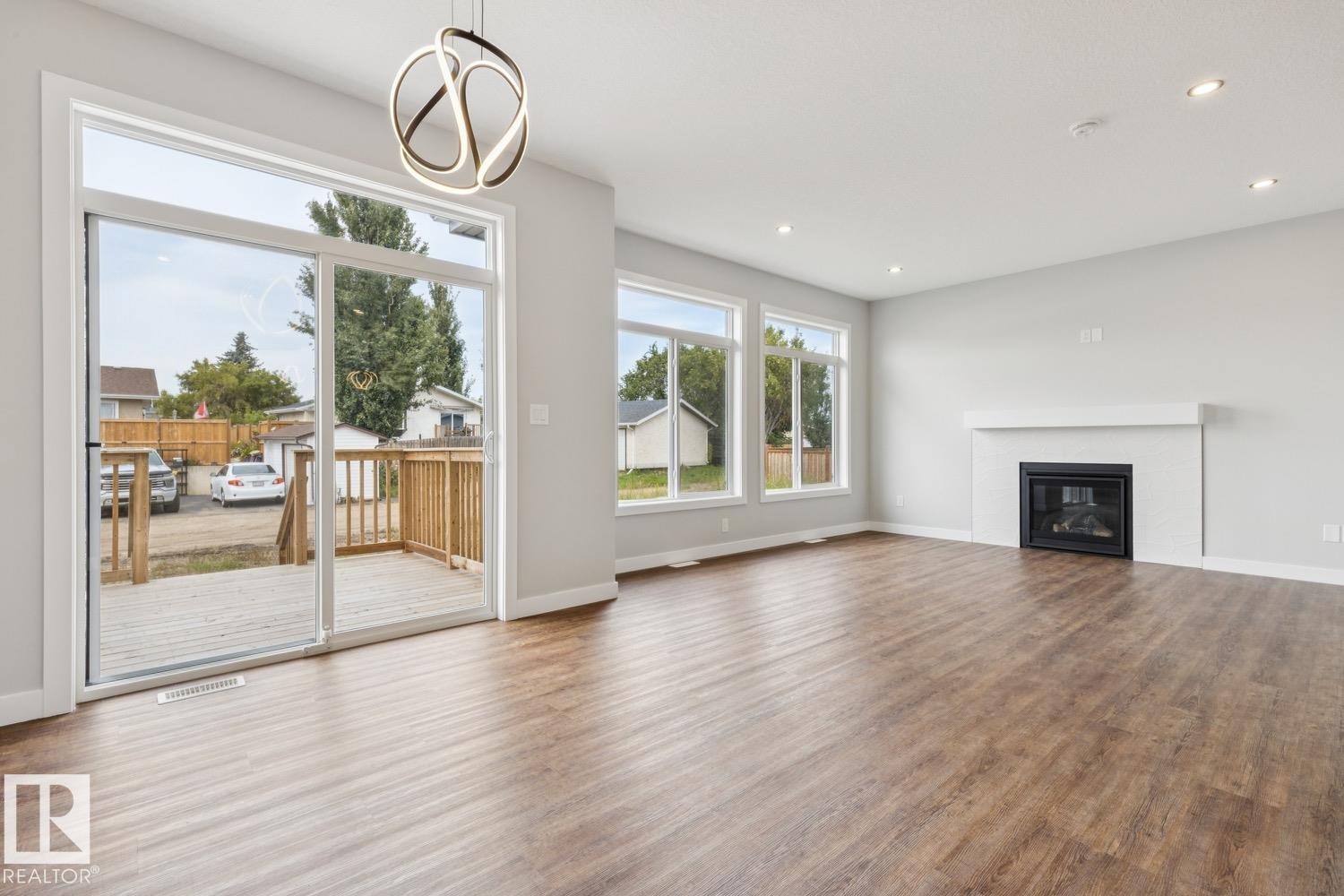 Spacious room featuring wood-finish flooring, a contemporary fireplace with a white mantel, multiple large windows, and a sliding glass door leading to a wooden deck - 72 Maple Crescent, Gibbons, AB
