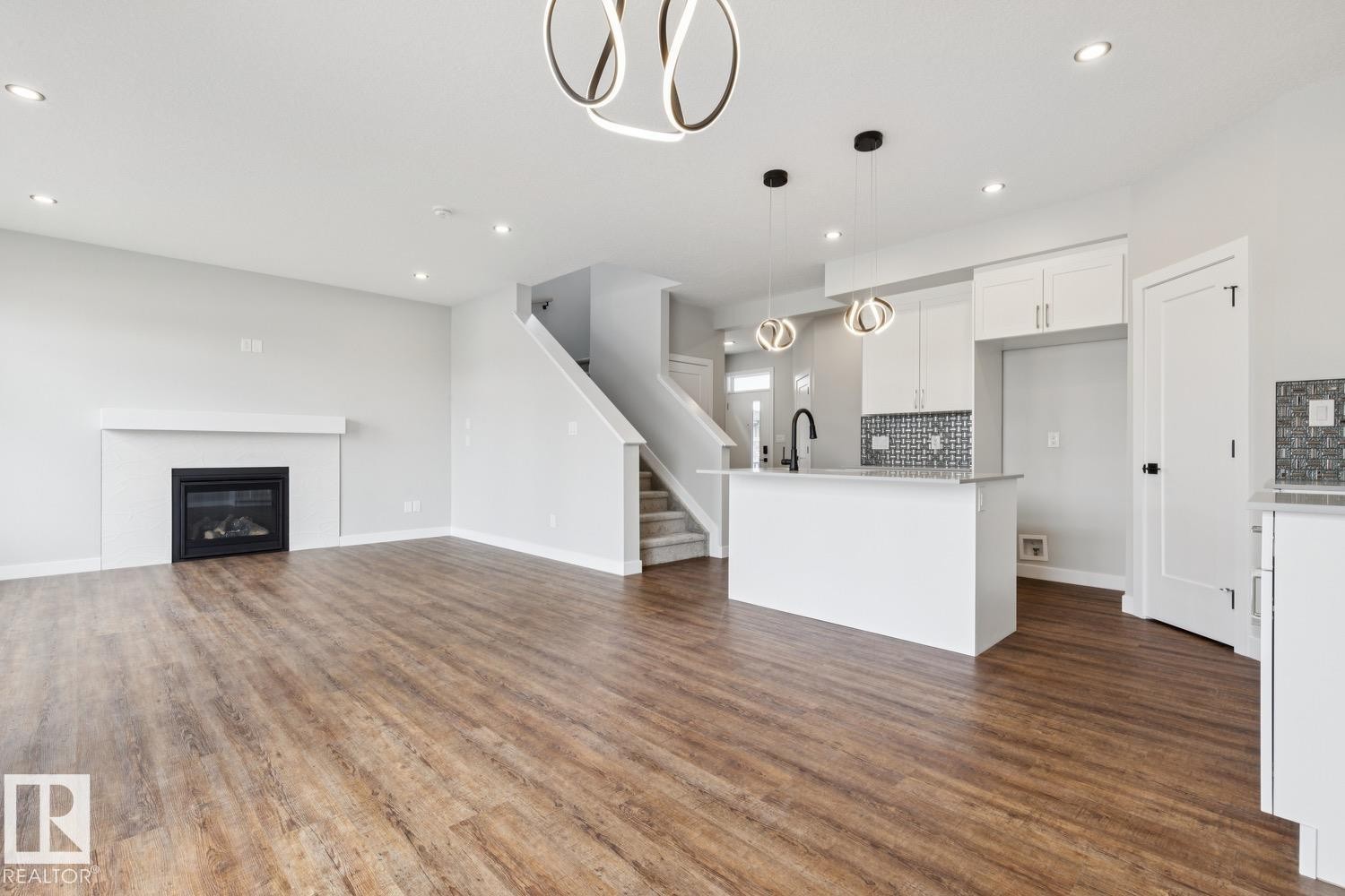 Wood-finish flooring, white kitchen cabinetry, a kitchen island with sink, a fireplace with a white mantel, and contemporary pendant lighting - 72 Maple Crescent, Gibbons, AB