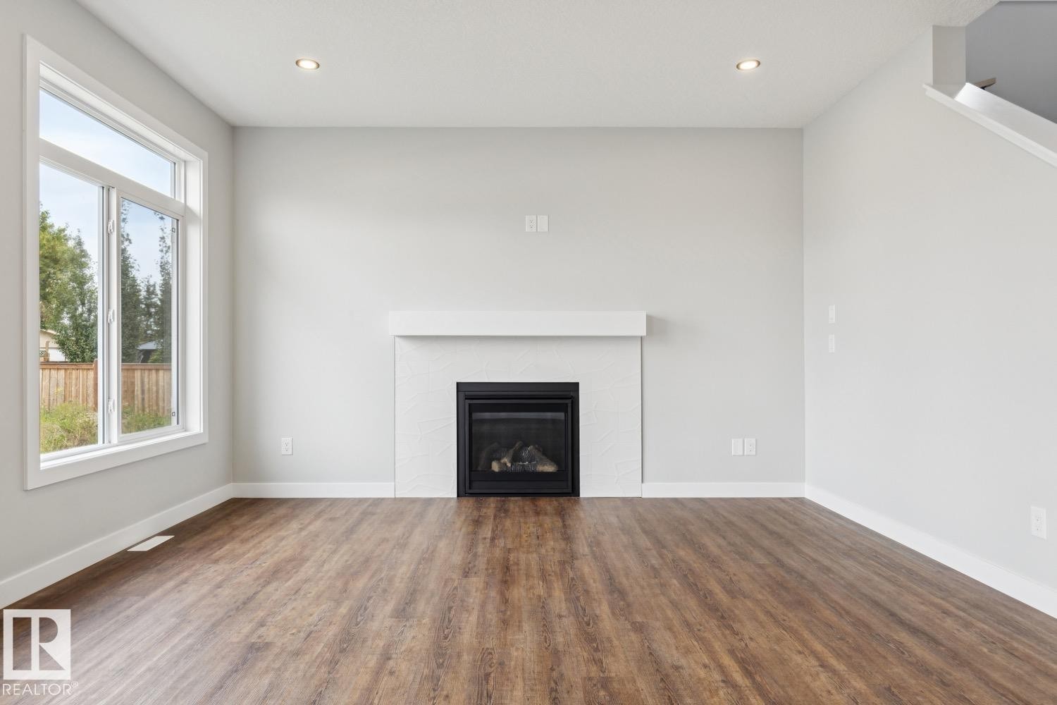 Modern fireplace with a white mantel, wood-finish flooring, recessed lighting, and a large window - 72 Maple Crescent, Gibbons, AB