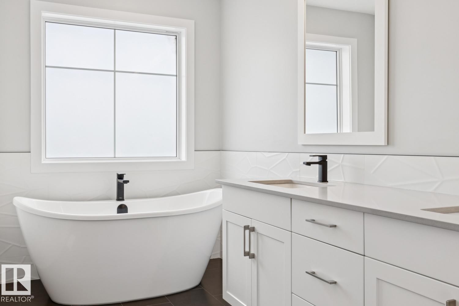 Freestanding soaking tub with a matte black faucet, double vanity with integrated sinks, white quartz countertop, and textured tile wainscoting - 72 Maple Crescent, Gibbons, AB