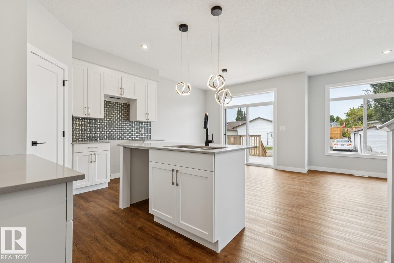 Modern kitchen featuring white cabinetry, a tiled backsplash, wood-finish flooring, and a kitchen island with a sink and pendant lighting - 72 Maple Crescent, Gibbons, AB