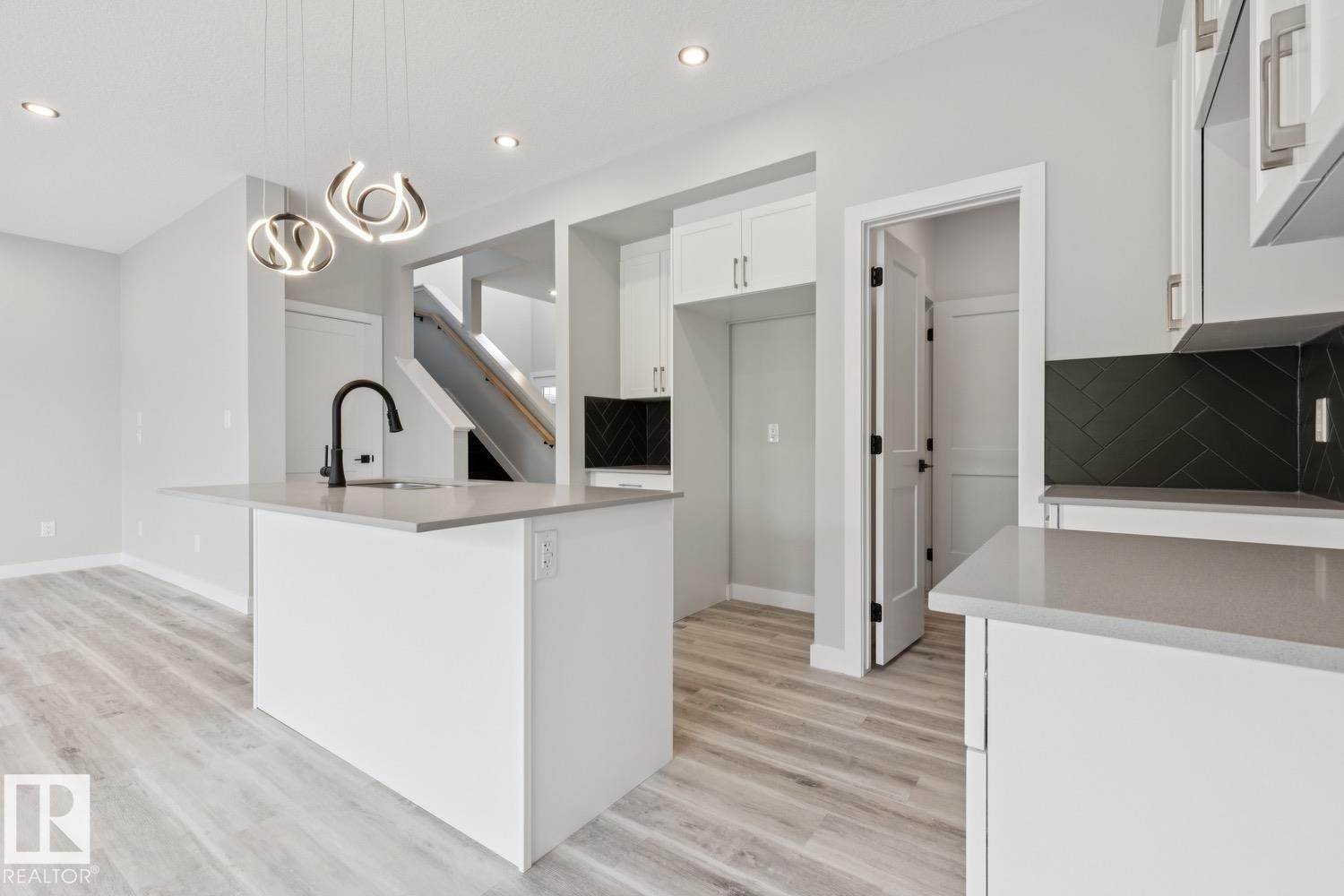 Kitchen featuring a central island with a sink, light-toned countertops, white cabinetry, a black herringbone tile backsplash, and wood-finish flooring - 36 Maple Crescent, Gibbons, AB