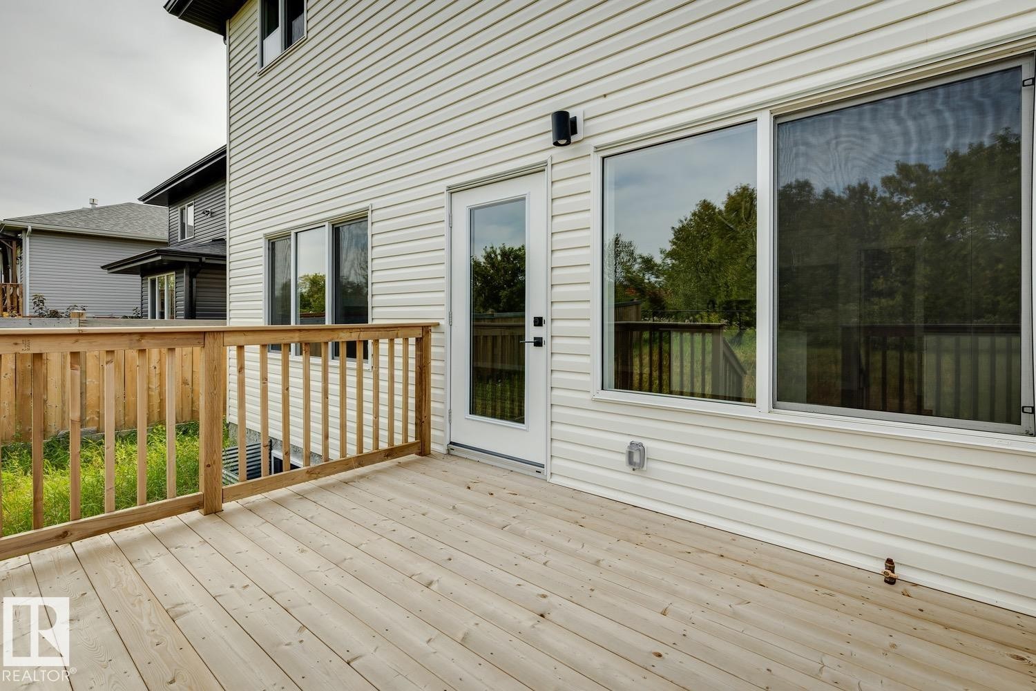 Expansive wood deck with a natural finish, featuring a full-light exterior door, large windows, and white vinyl siding - 36 Maple Crescent, Gibbons, AB