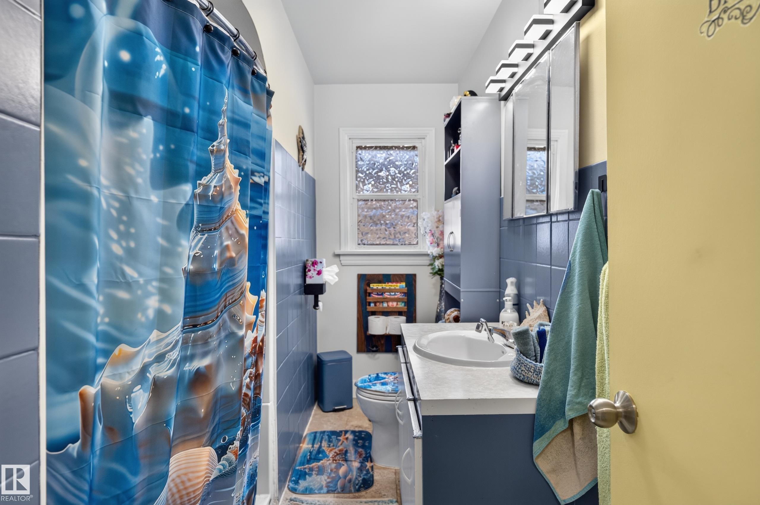 Full bathroom featuring a single vanity with an integrated sink, blue tile wainscoting, and a window with privacy glass - 12309 80 Street Nw, Edmonton, AB