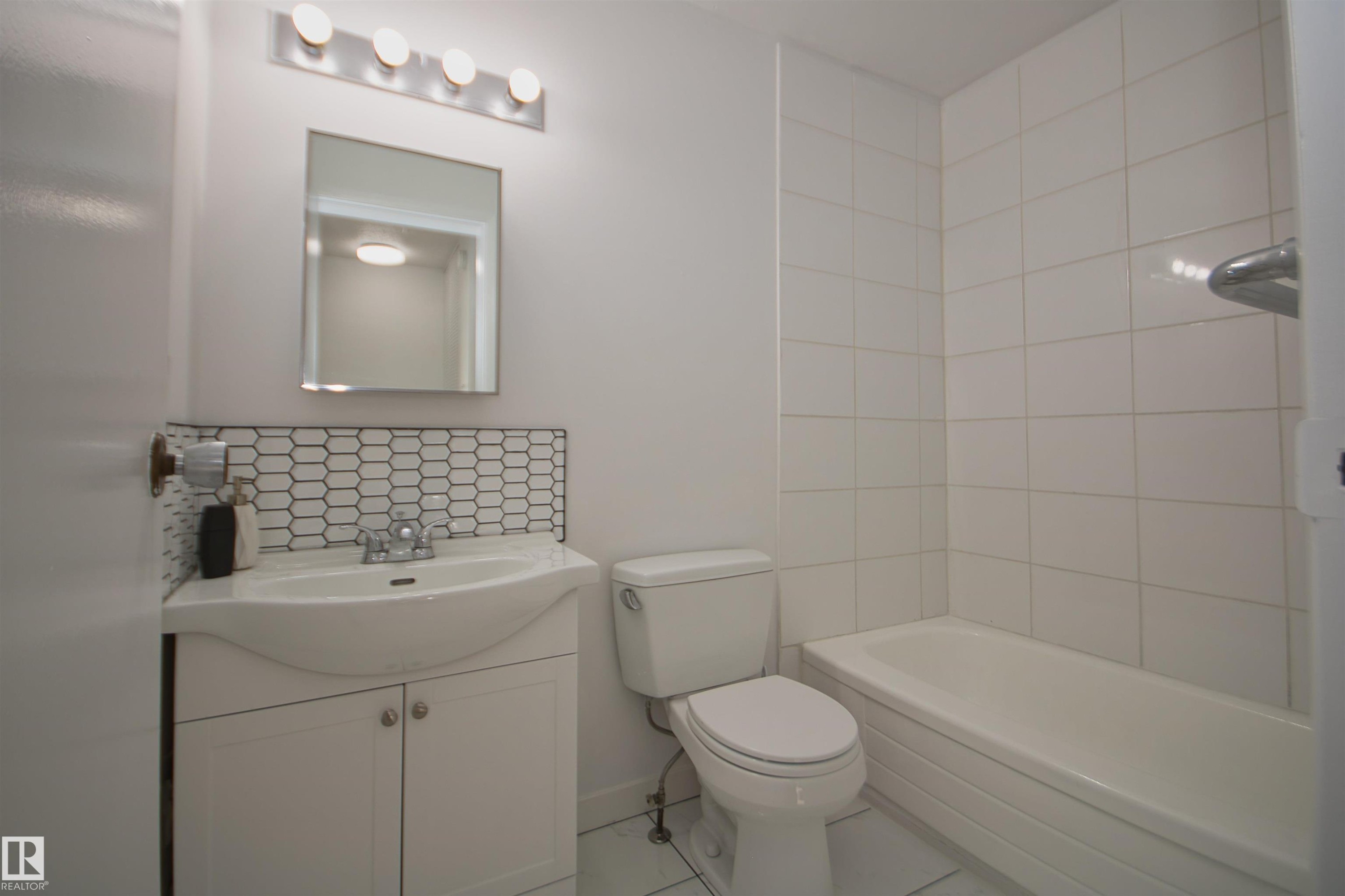 White subway tile shower surround, integrated bathtub, white vanity with storage, mosaic tile backsplash, and wall-mounted vanity lighting - 7743 37 Avenue Nw, Edmonton, AB