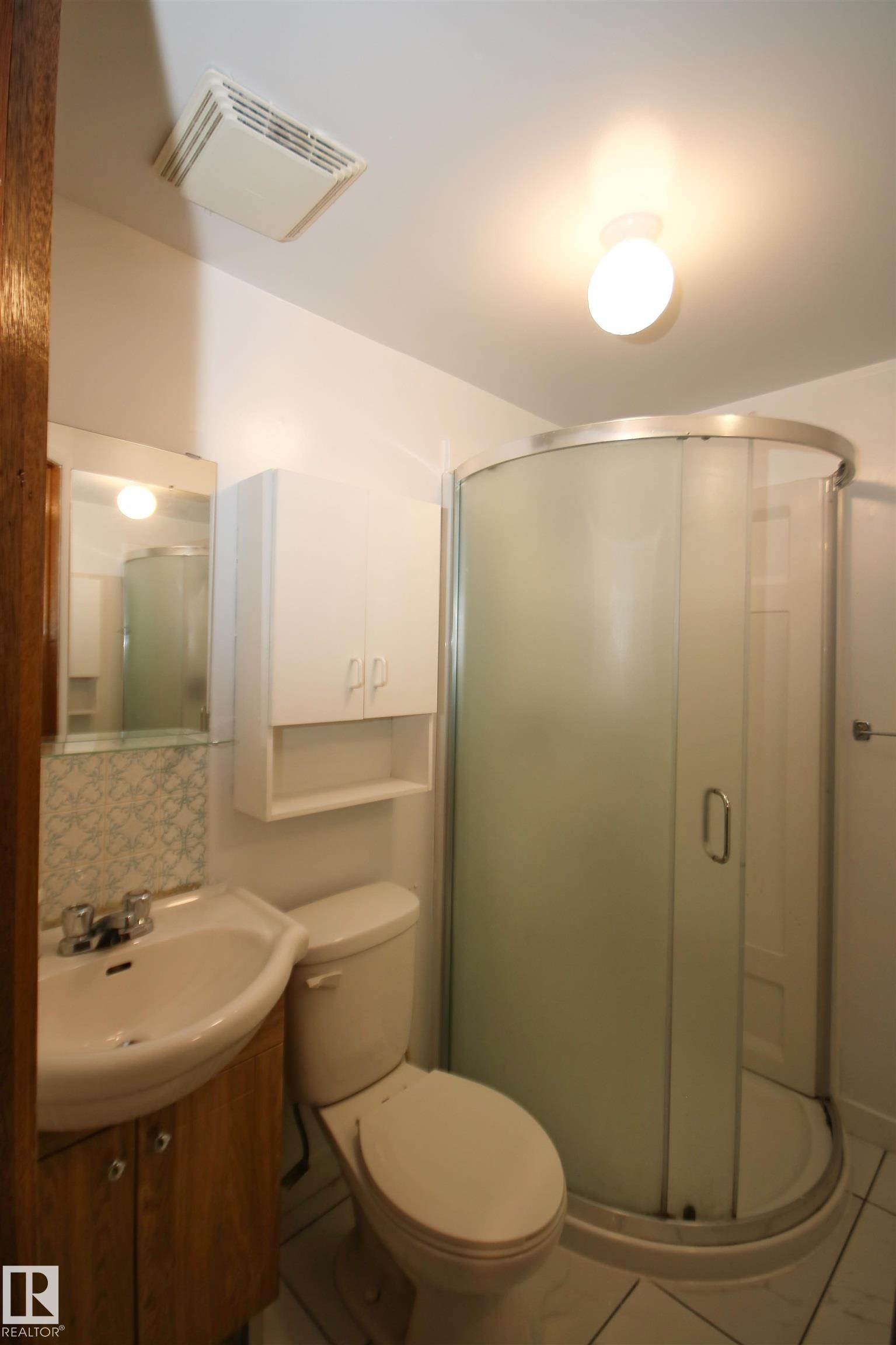 Compact bathroom featuring a curved glass shower enclosure, white wall-mounted storage cabinet, and a pedestal sink with a wood-finish vanity - 7743 37 Avenue Nw, Edmonton, AB