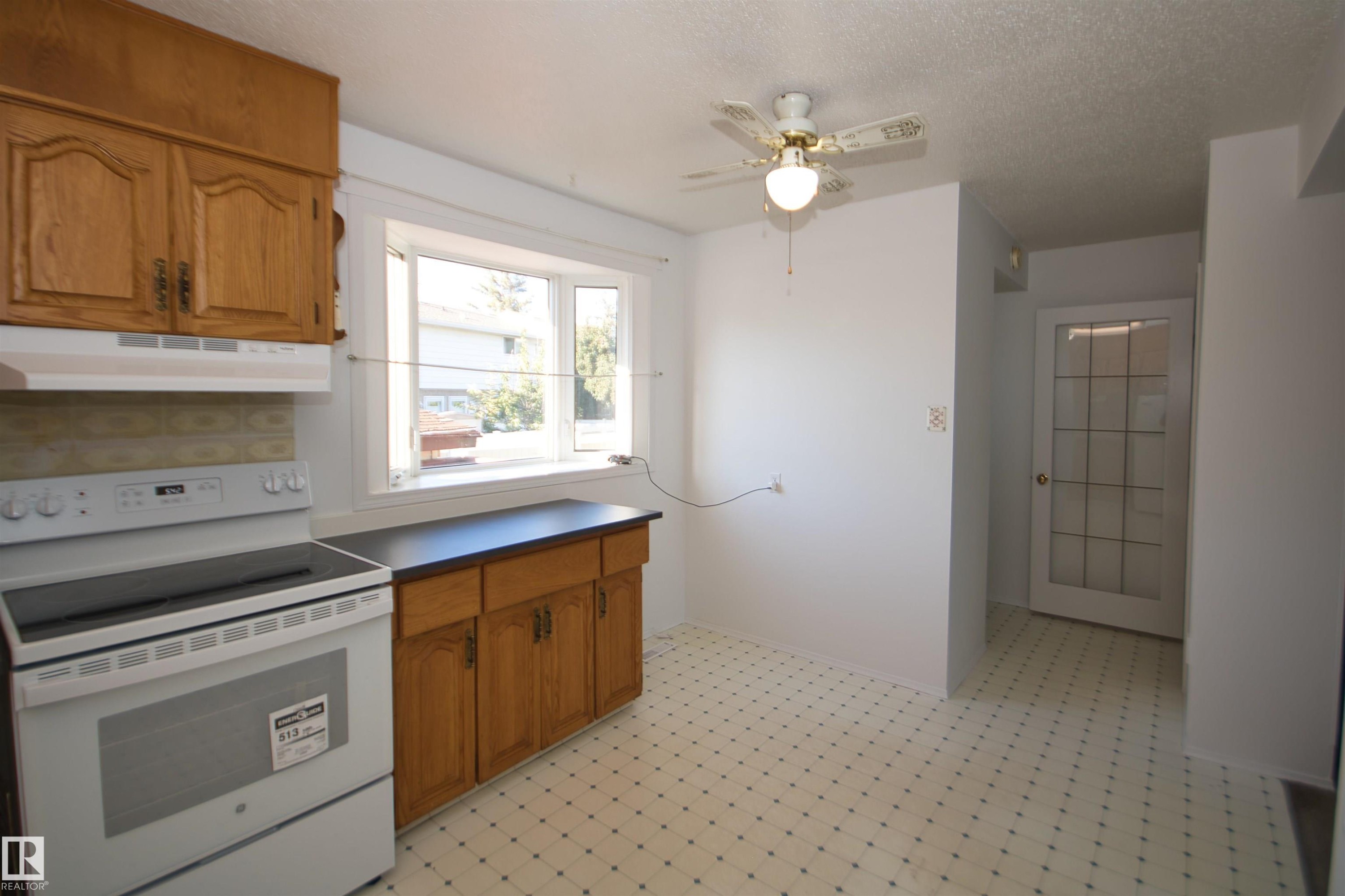Wood cabinetry with dark countertops, white electric range, bay window, ceiling fan with light fixture, and a glass-paneled door - 7743 37 Avenue Nw, Edmonton, AB