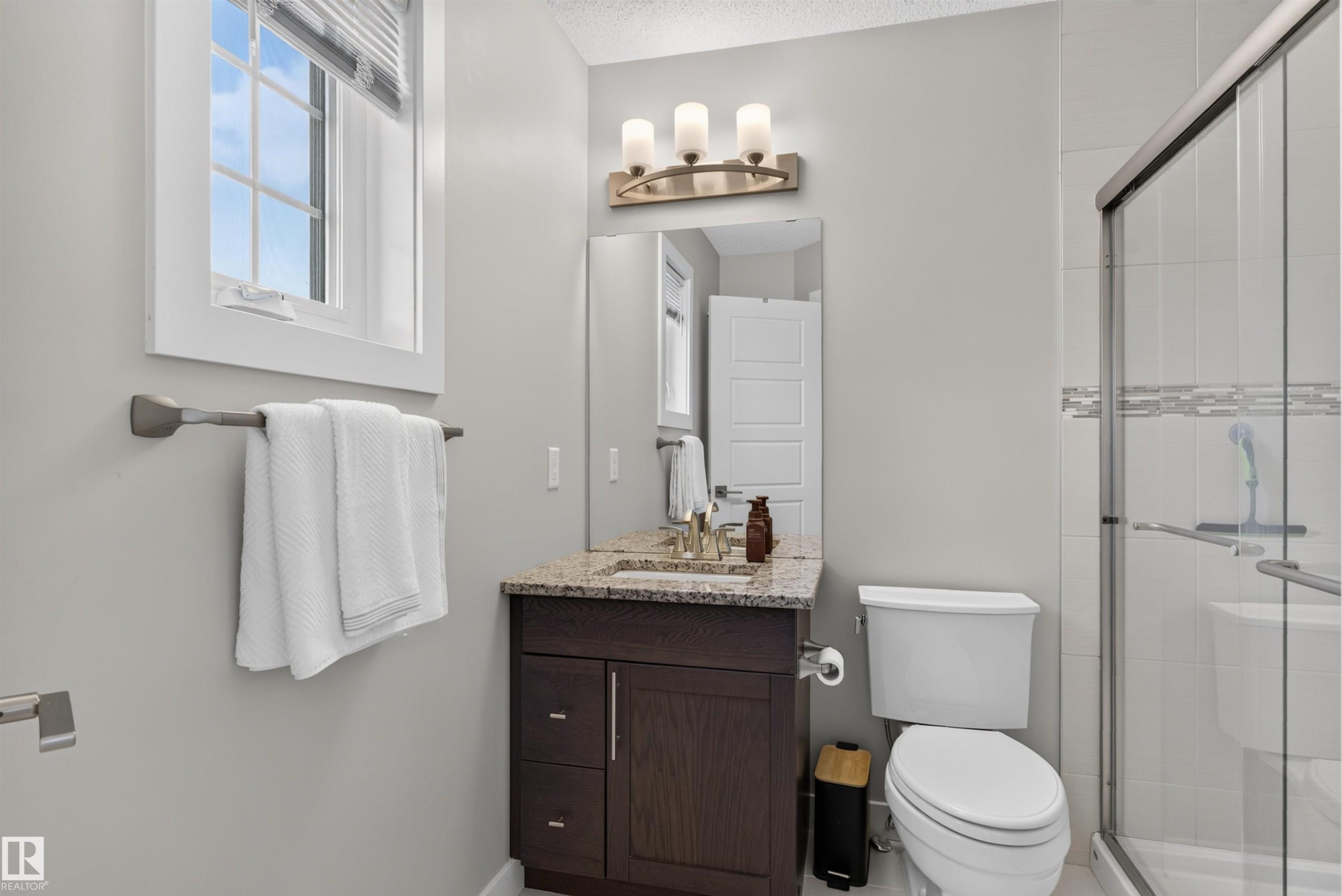 Wood-finish vanity with a stone countertop, framed mirror, and three-light fixture - 319 Desrochers Boulevard Sw, Edmonton, AB